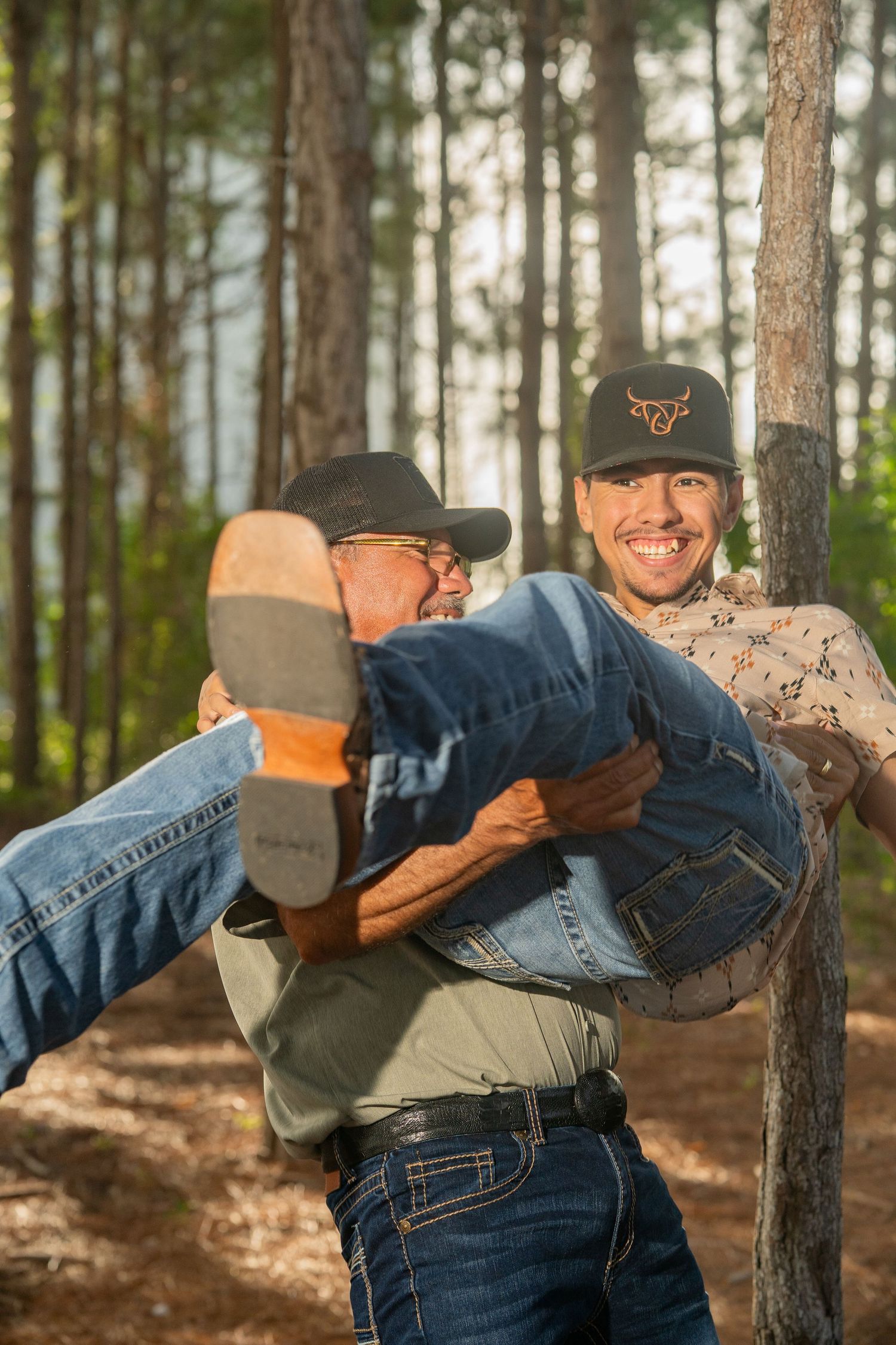 Two men in cowboy hats laugh in a pine forest, one carrying the other horizontally in his arms.