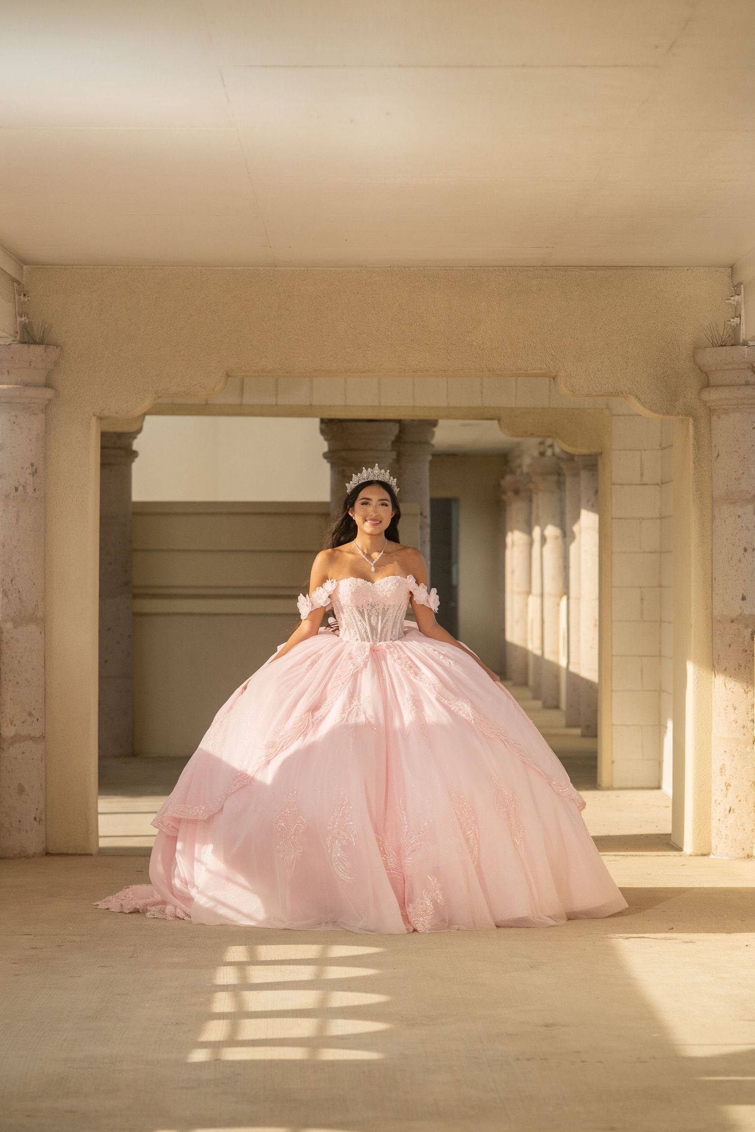 Young woman in a pink ball gown and tiara poses in a sunlit stone colonnade for her quinceañera.