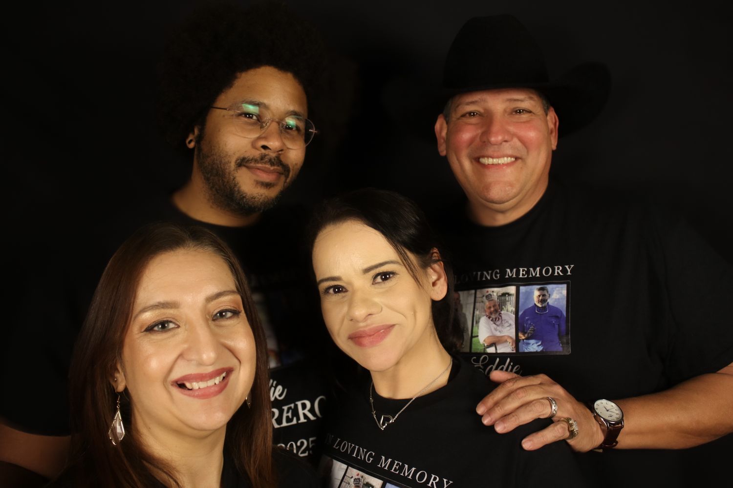 A group of four people wearing black t-shirts smile together in a casual photo against a dark background.