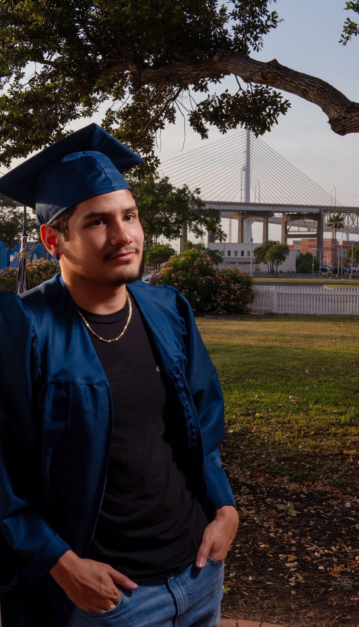 A graduate in blue cap and gown stands thoughtfully near a tree with a bridge visible in the background.