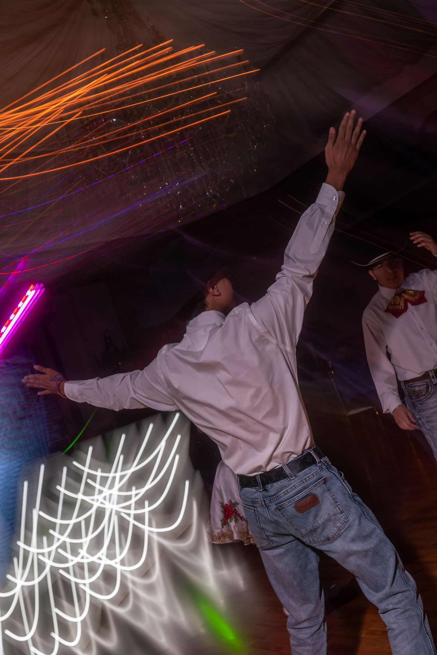 Dancer in white shirt and jeans performs energetic move under colorful light streaks at nightclub.