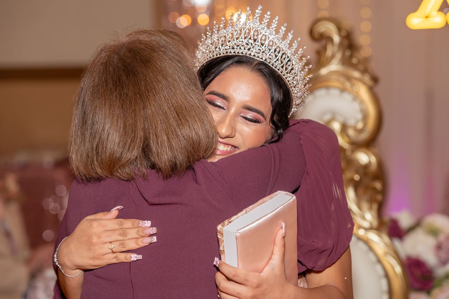 A quinceañera girl wearing a crystal tiara shares a warm embrace with a guest at her celebration.