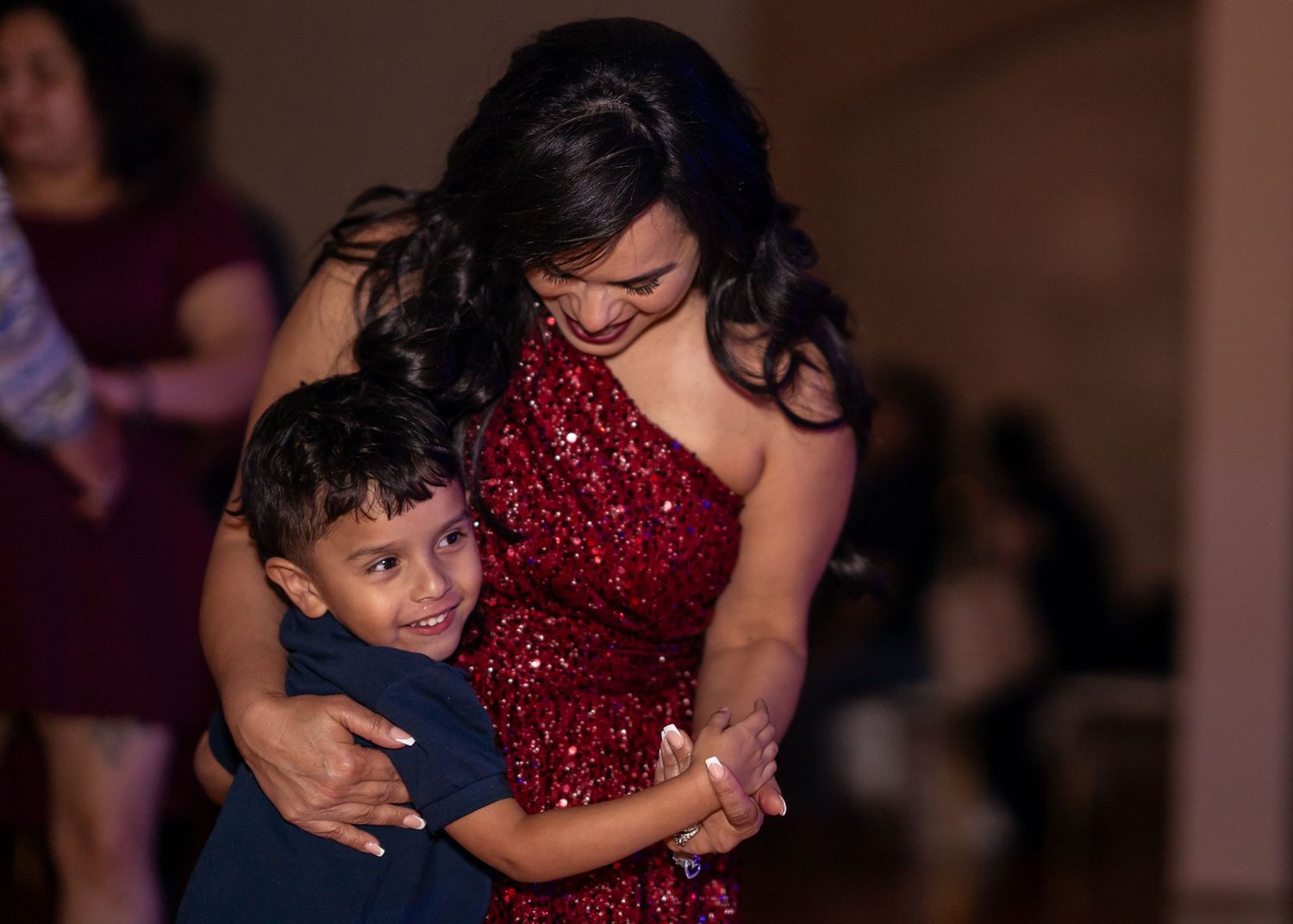A woman in a red sequin dress hugs a young boy on the dance floor at a festive celebration.