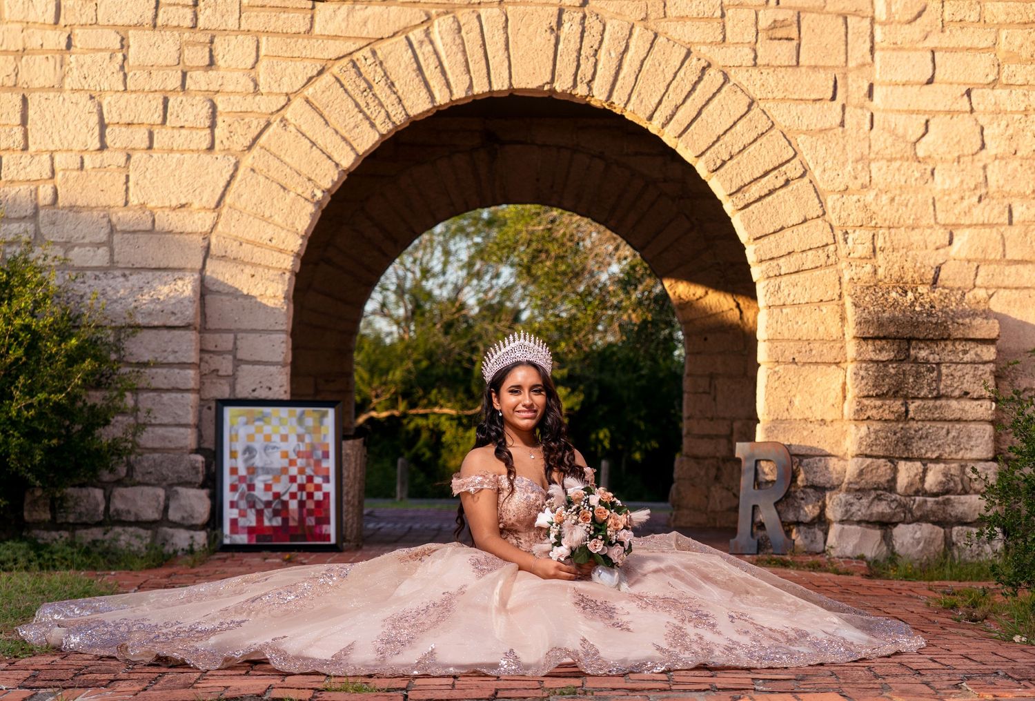A bride in a blush pink gown sits before a stone archway at sunset holding a floral bouquet.