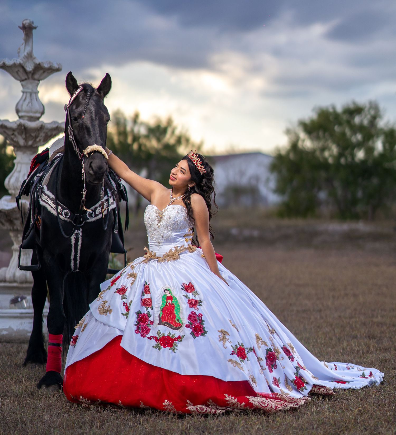 Elegant white ball gown with red floral embroidery photographed outdoors with a black horse at dusk.