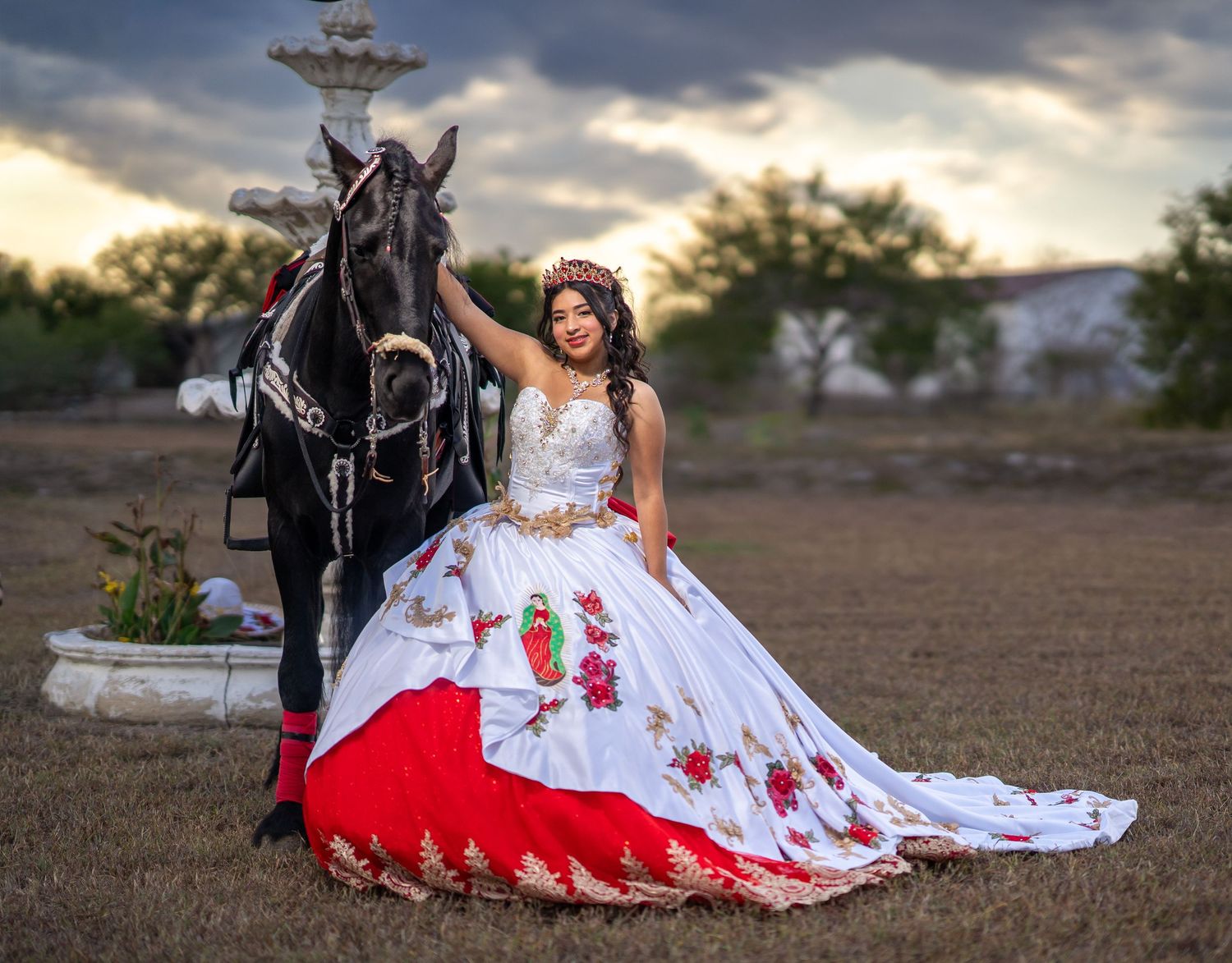 Traditional Mexican quinceañera dress with red and white embroidery details shown alongside a black horse at sunset.