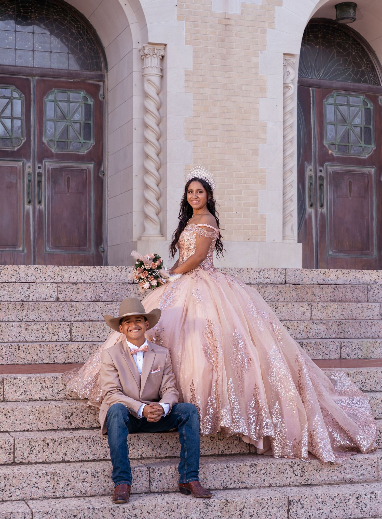 A couple in contrasting formal wear sits together on stone steps with ornate architectural details behind them.