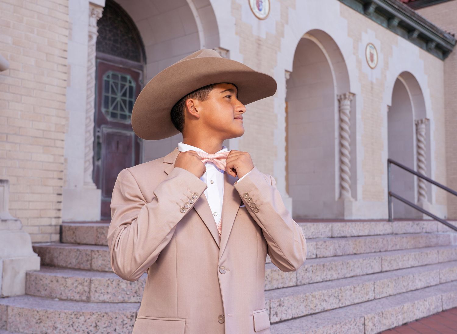 A person wearing a beige blazer and cowboy hat stands against a light brick building with arched windows.