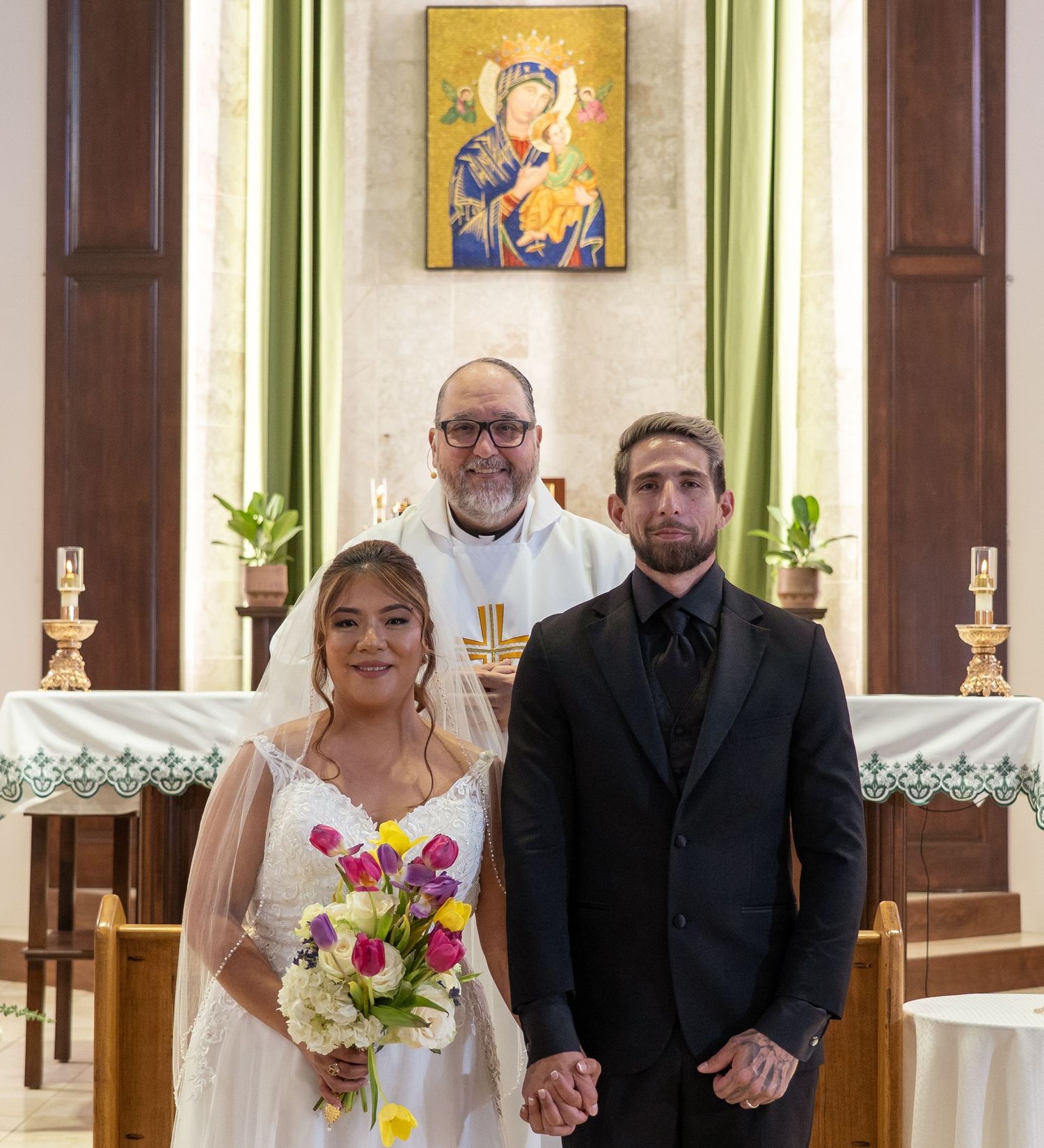 Wedding party stands in formal attire at the altar of a traditional church with religious artwork on walls.