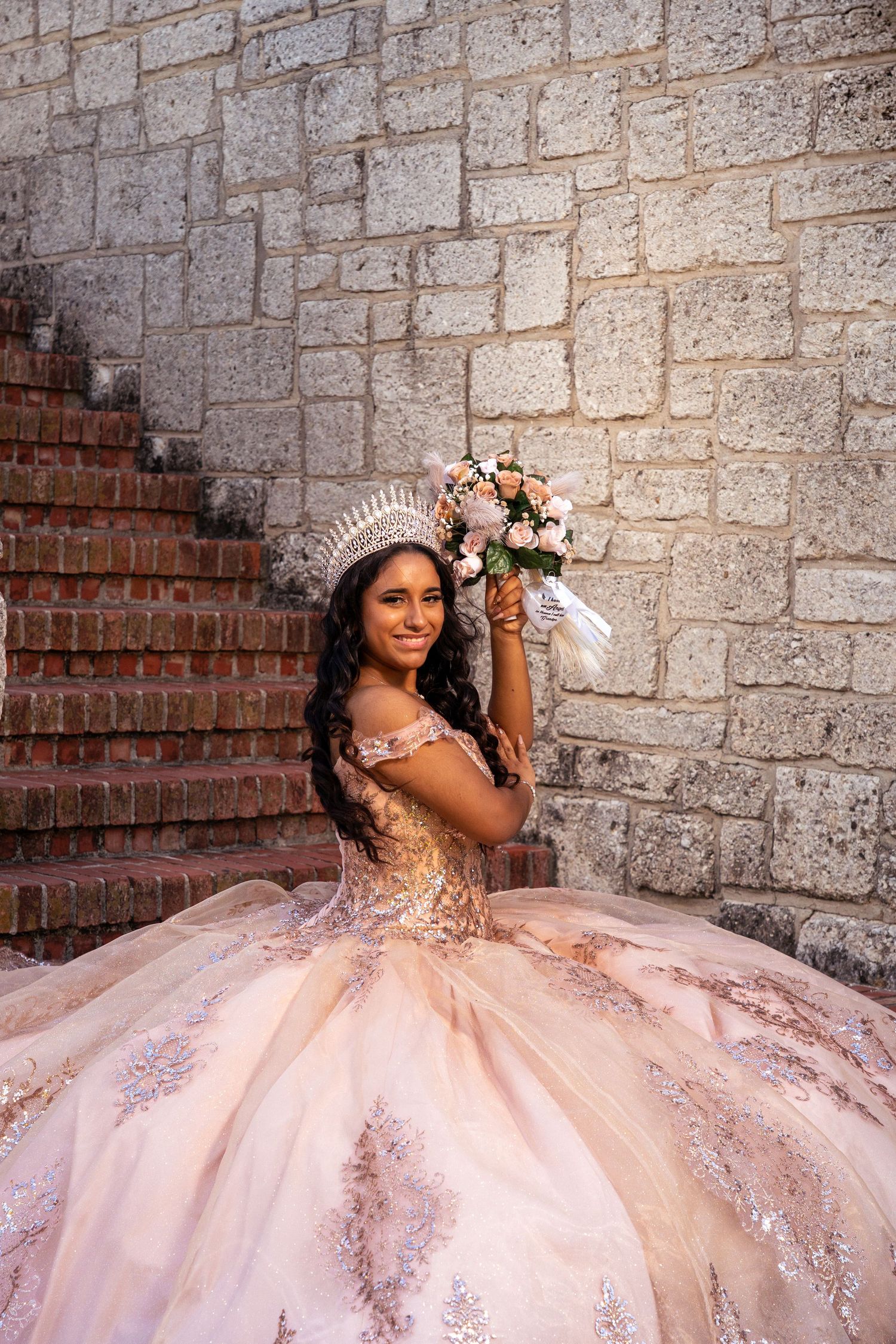 A person in a pink quinceañera ball gown with sparkly details poses on stone steps next to a brick wall.