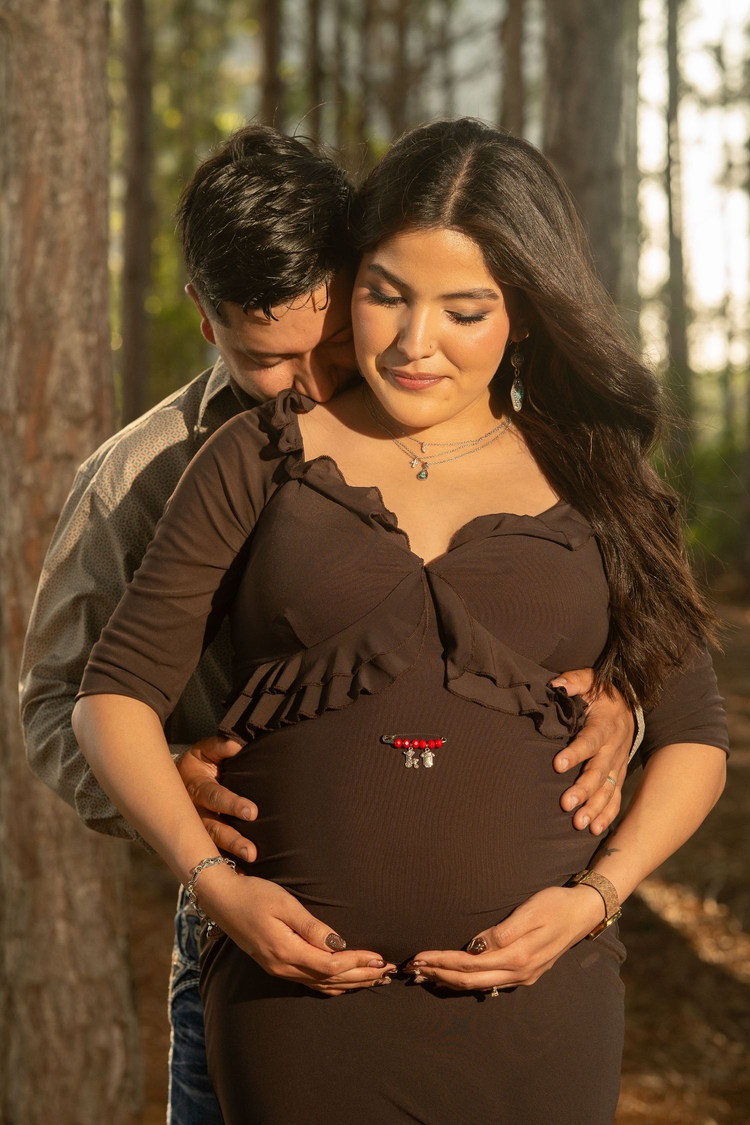 Couple in forest maternity photo, woman in brown dress cradling baby bump, man embracing her from behind.