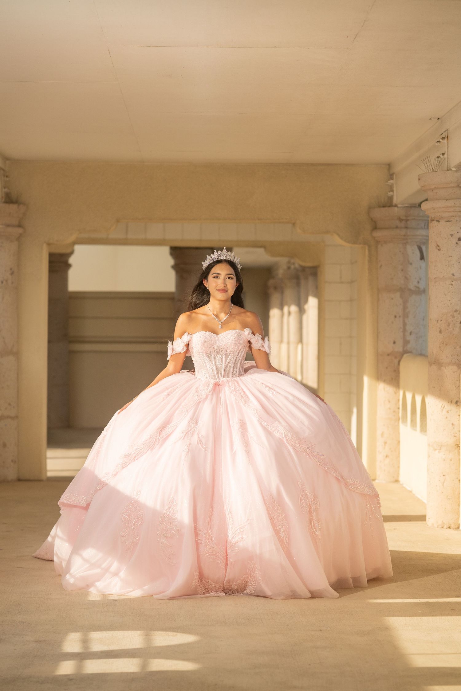 Young woman in pink ball gown and tiara poses gracefully in sunlit stone archway for quinceañera.