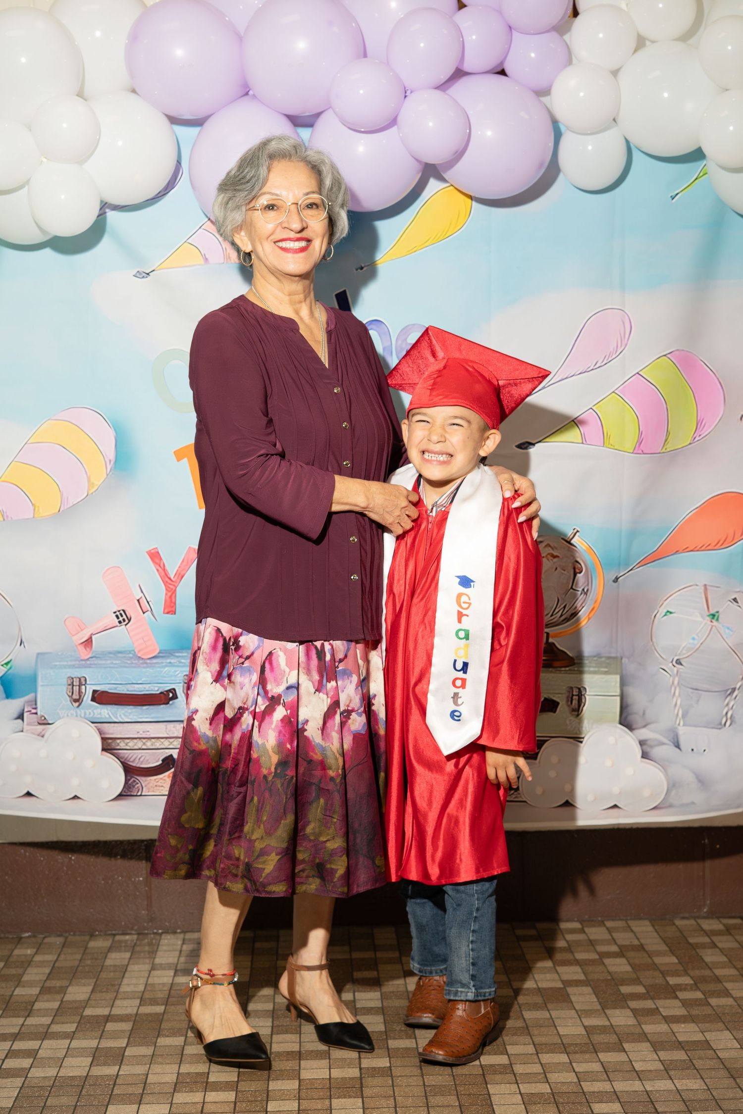 Grandmother proudly poses with young child in red graduation cap and gown at kindergarten graduation ceremony.