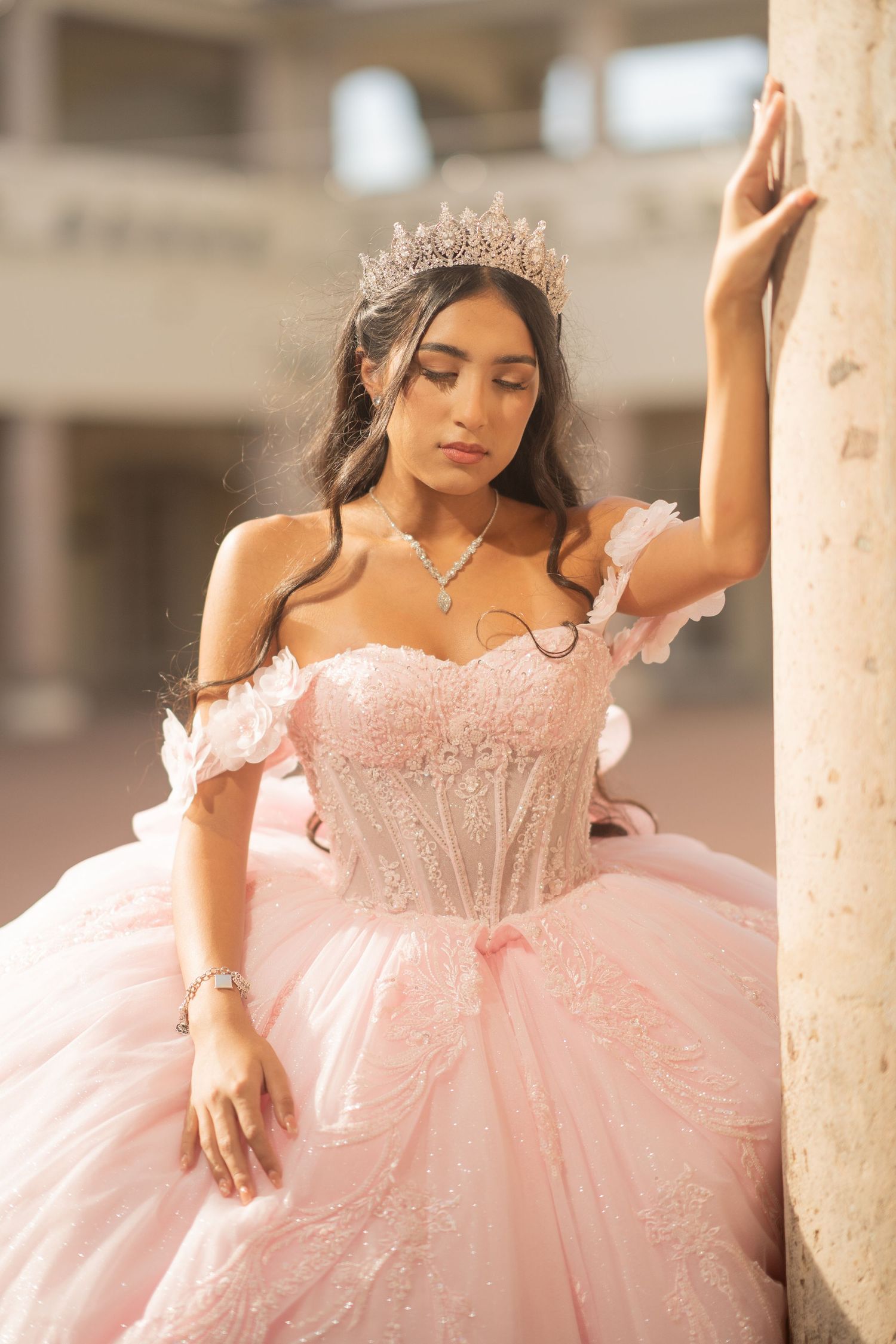 Young woman in pink ball gown and crystal tiara poses gracefully against a column for quinceañera photos.