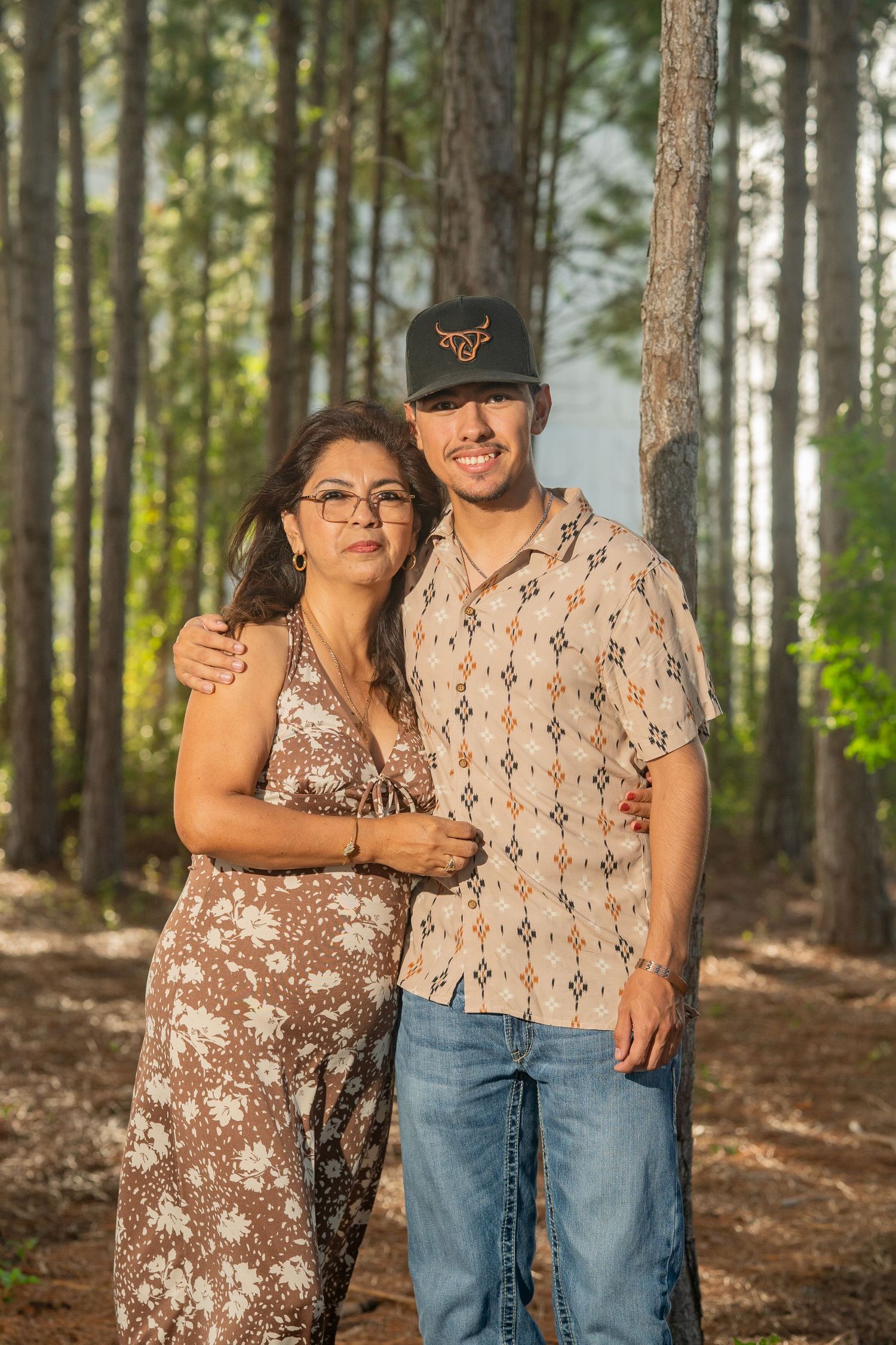 A woman and young man pose together in a sunlit pine forest, she in a floral dress, he in a patterned shirt and cap.