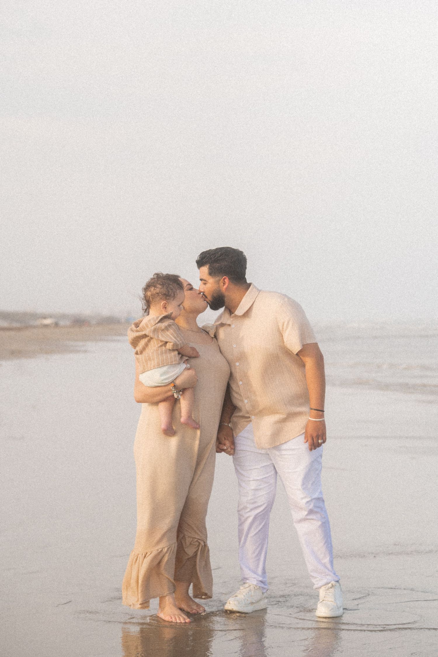 A loving couple shares a kiss on a misty beach while the woman holds their young baby in her arms.
