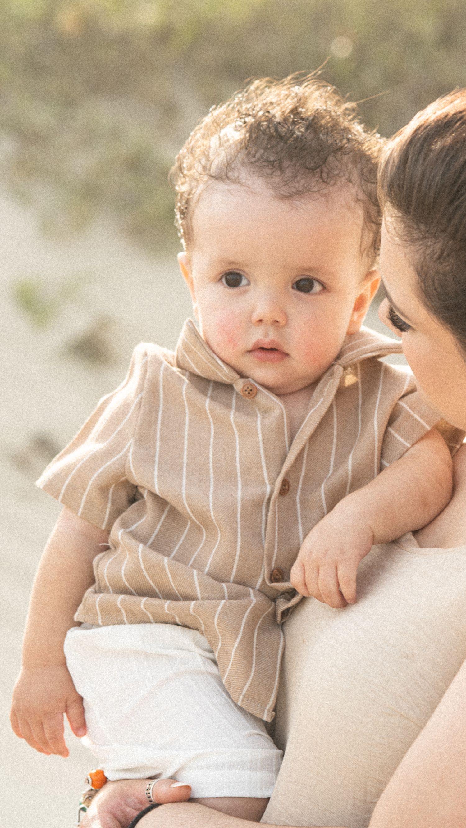 Adorable baby in striped linen shirt held by mother on sunny beach.
