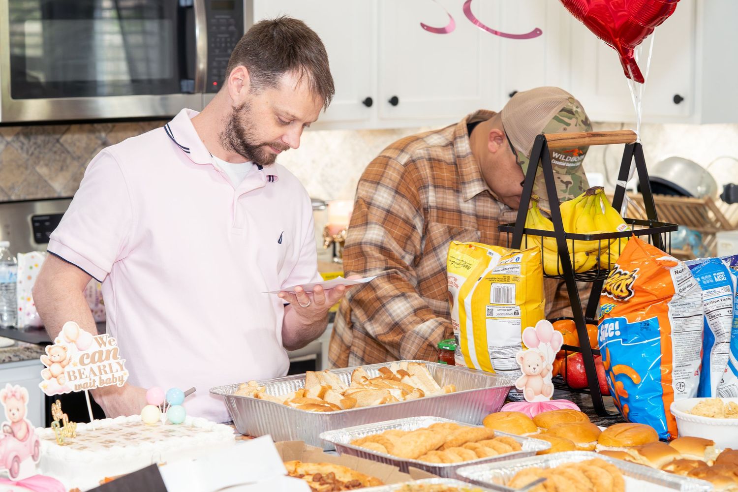 Two men serving food at a baby shower buffet table with snacks, pastries, and festive decorations in a kitchen.