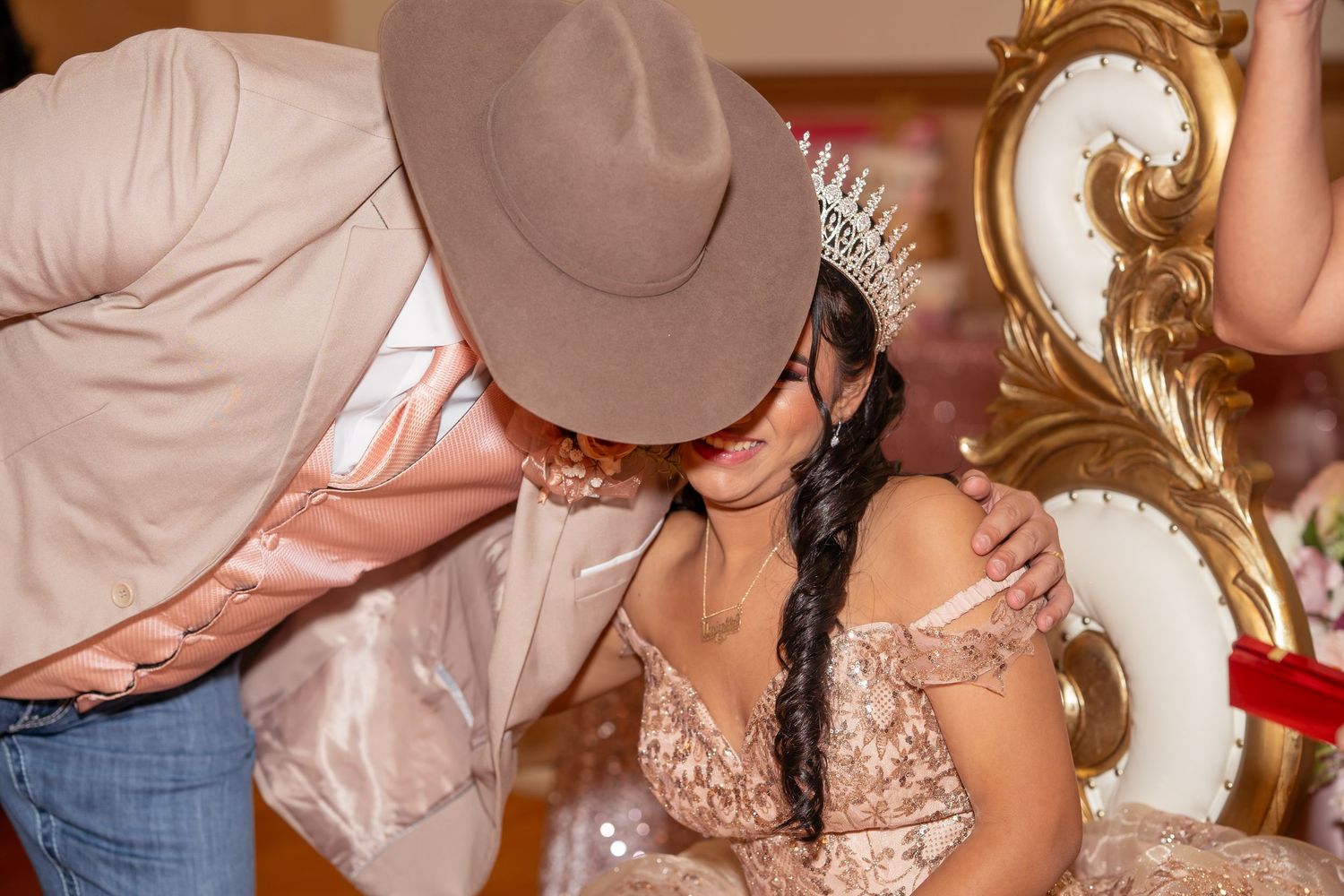 A man in a cowboy hat leans down to embrace a young woman in a rose gold Quinceañera gown seated on a throne.