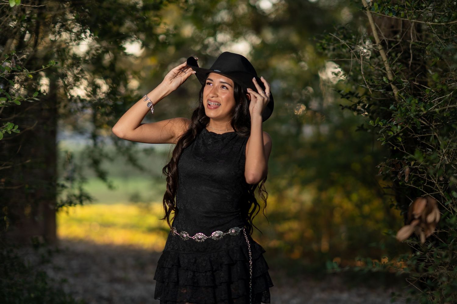A young woman in a black dress and hat poses gracefully among lush green trees in an outdoor setting.