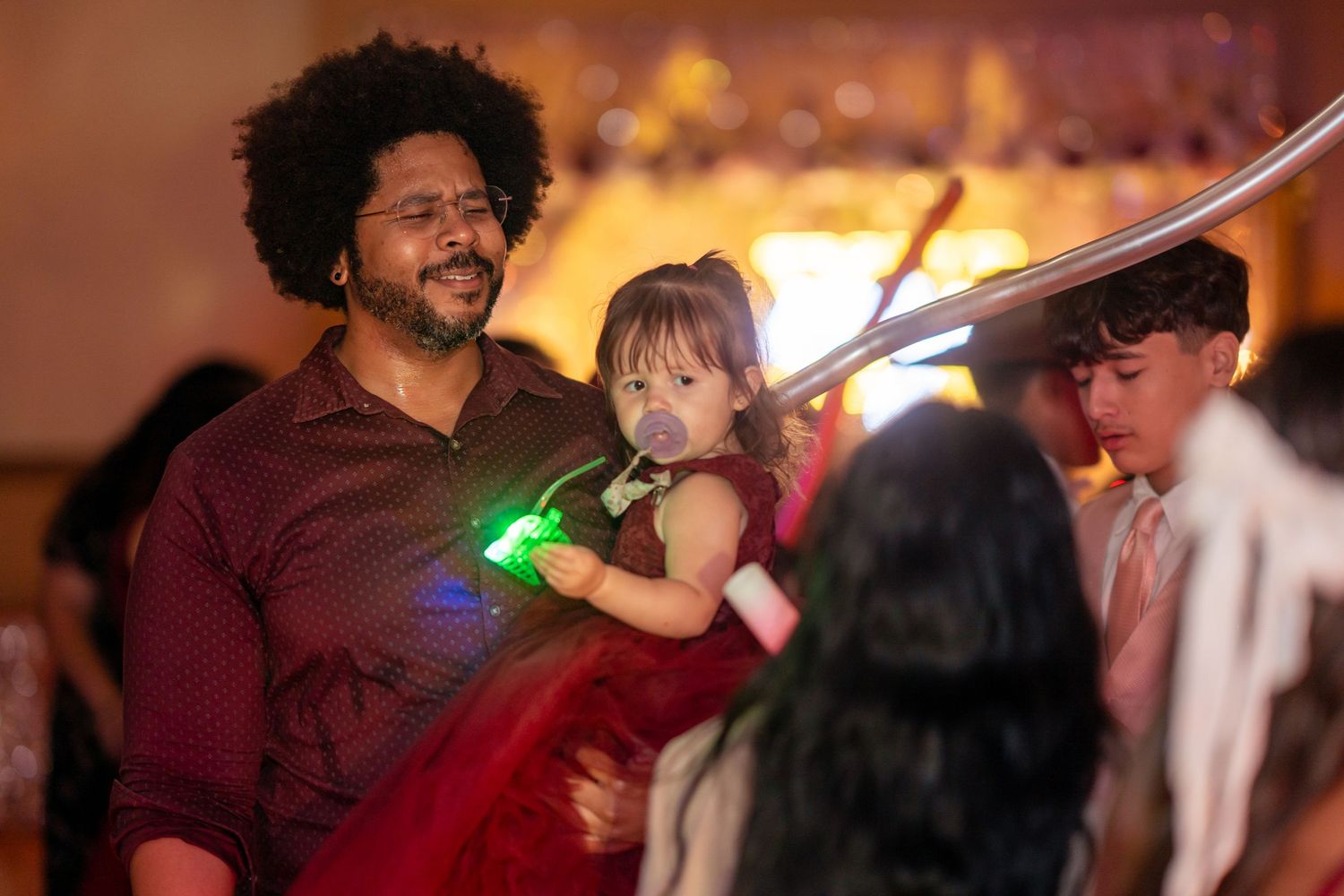 A man with an afro holds a toddler girl in a red dress while glowing accessories light up a party dance floor.