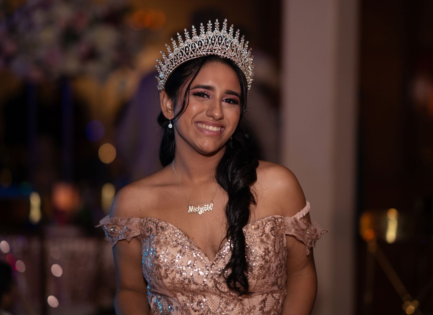 Young woman in a rose gold off-shoulder gown and diamond tiara smiles radiantly at her quinceañera celebration.