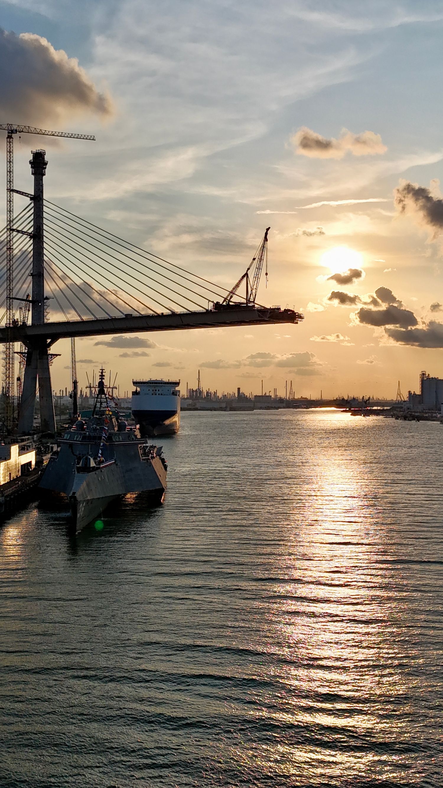 Ships and cranes silhouetted against a golden sunset at a busy port with a cable-stayed bridge in the background.