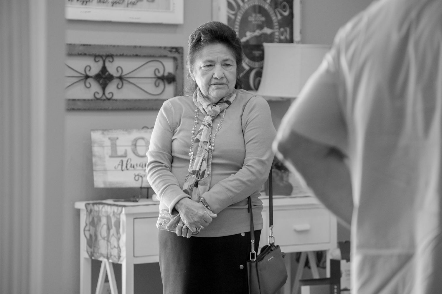 Black and white photo of an elderly Asian woman standing indoors, looking pensively at someone nearby.