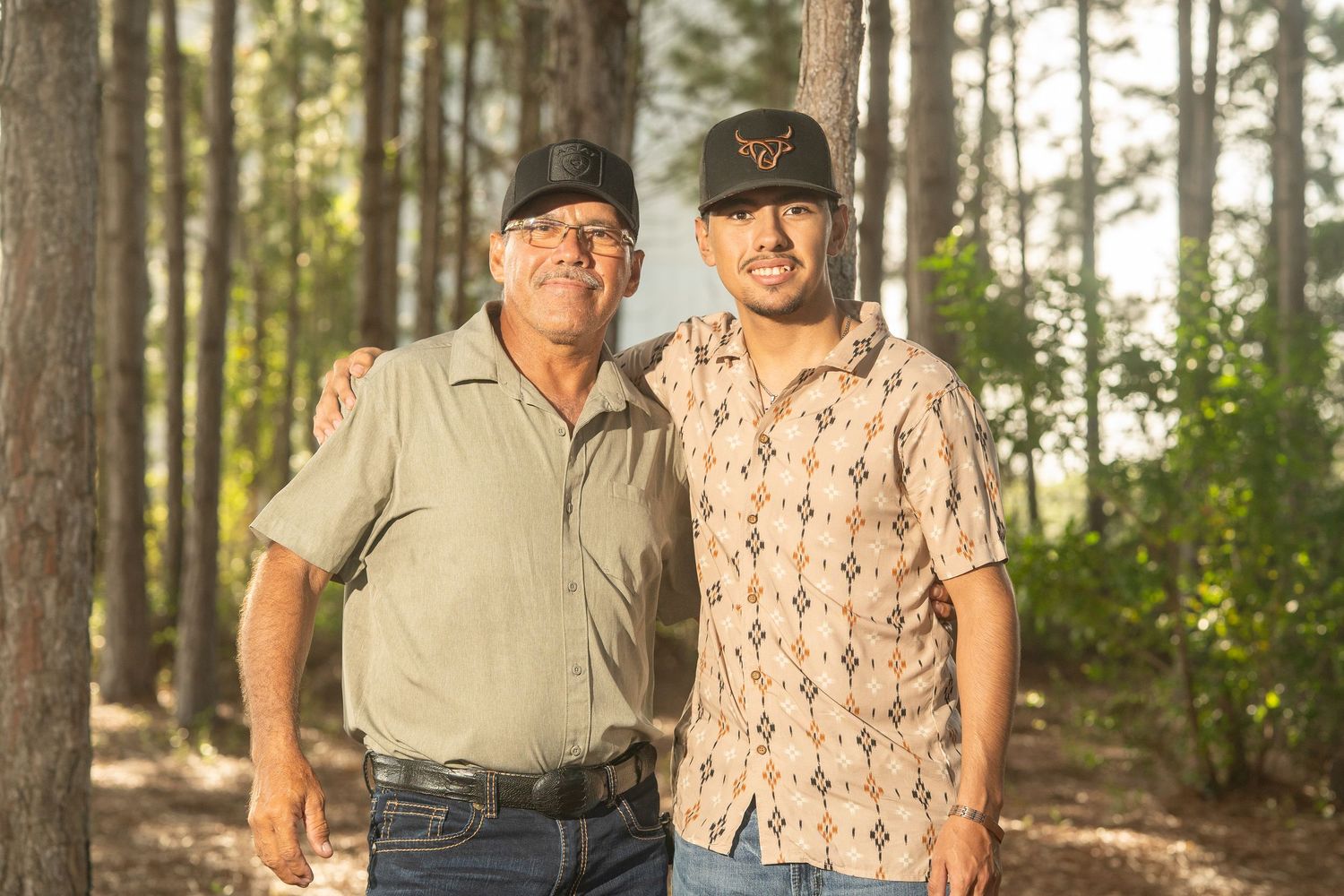 Two men wearing baseball caps pose together in a pine forest, smiling for a portrait photo outdoors.