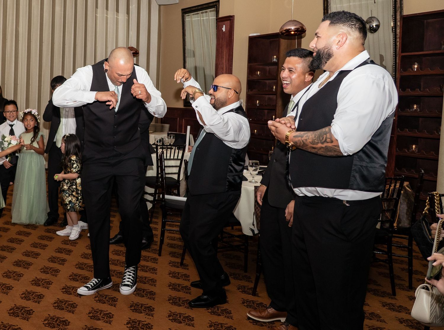 Groomsmen in black vests and white shirts dancing and celebrating joyfully at an elegant wedding reception.