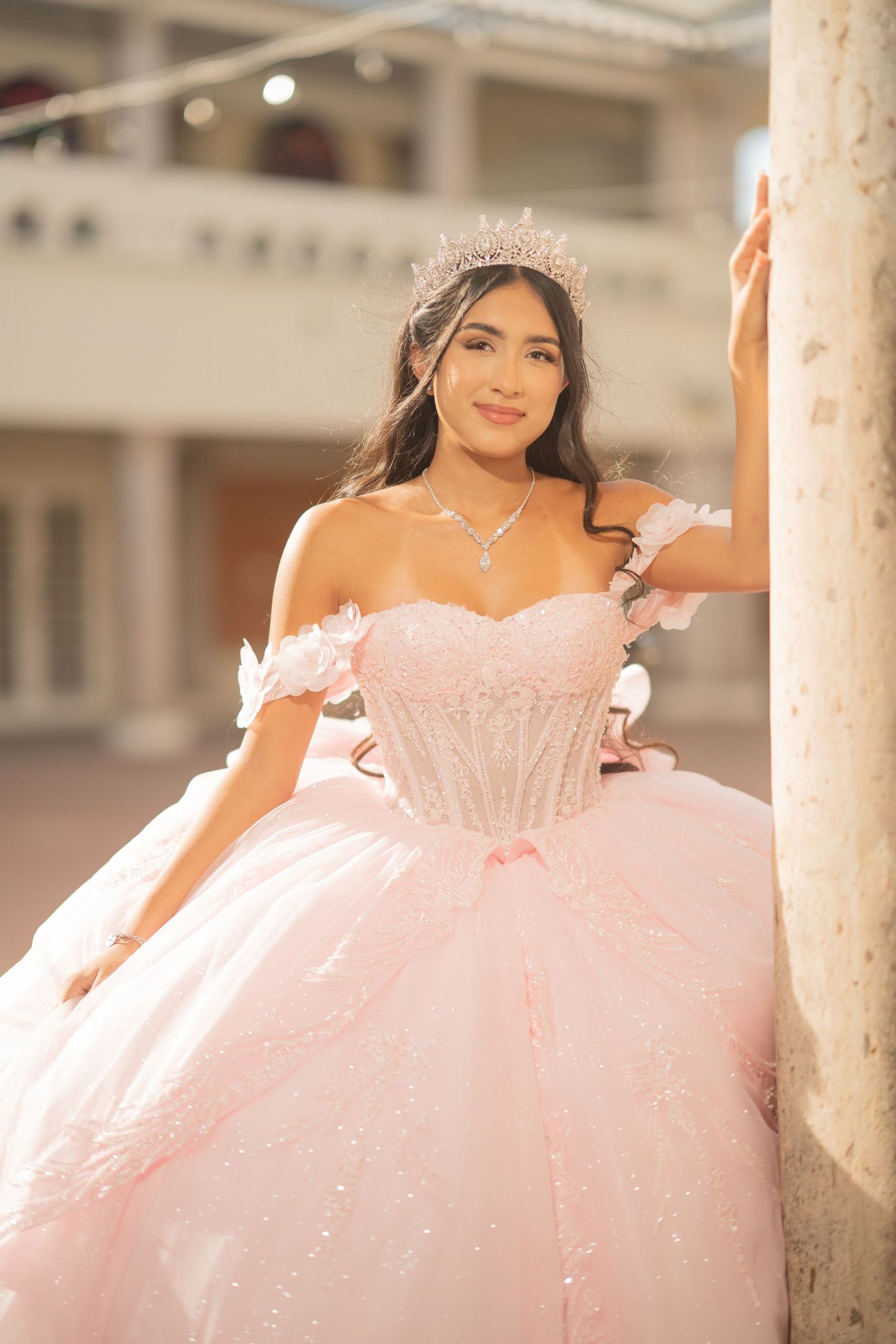 Young woman in pink quinceañera ball gown with tiara and off-shoulder floral details poses by a column outdoors.