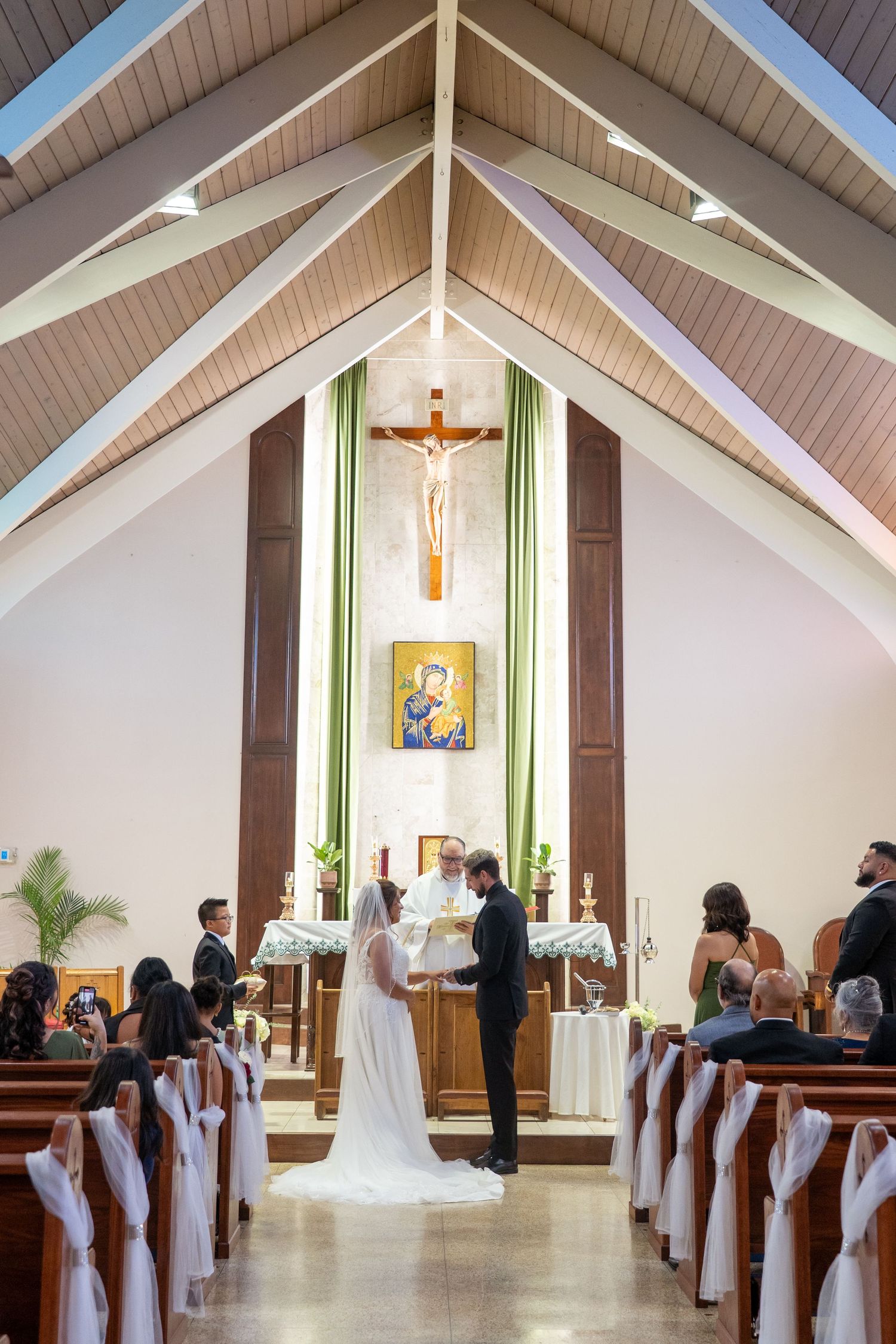 Modern church interior with vaulted white ceiling, wooden beams, and religious artwork during a wedding ceremony.