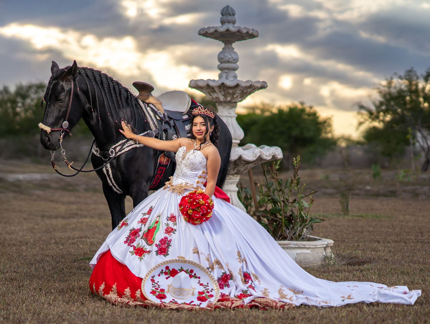 A dramatic sunset photo featuring a red and white dress, red roses, and a black horse near a stone cross.
