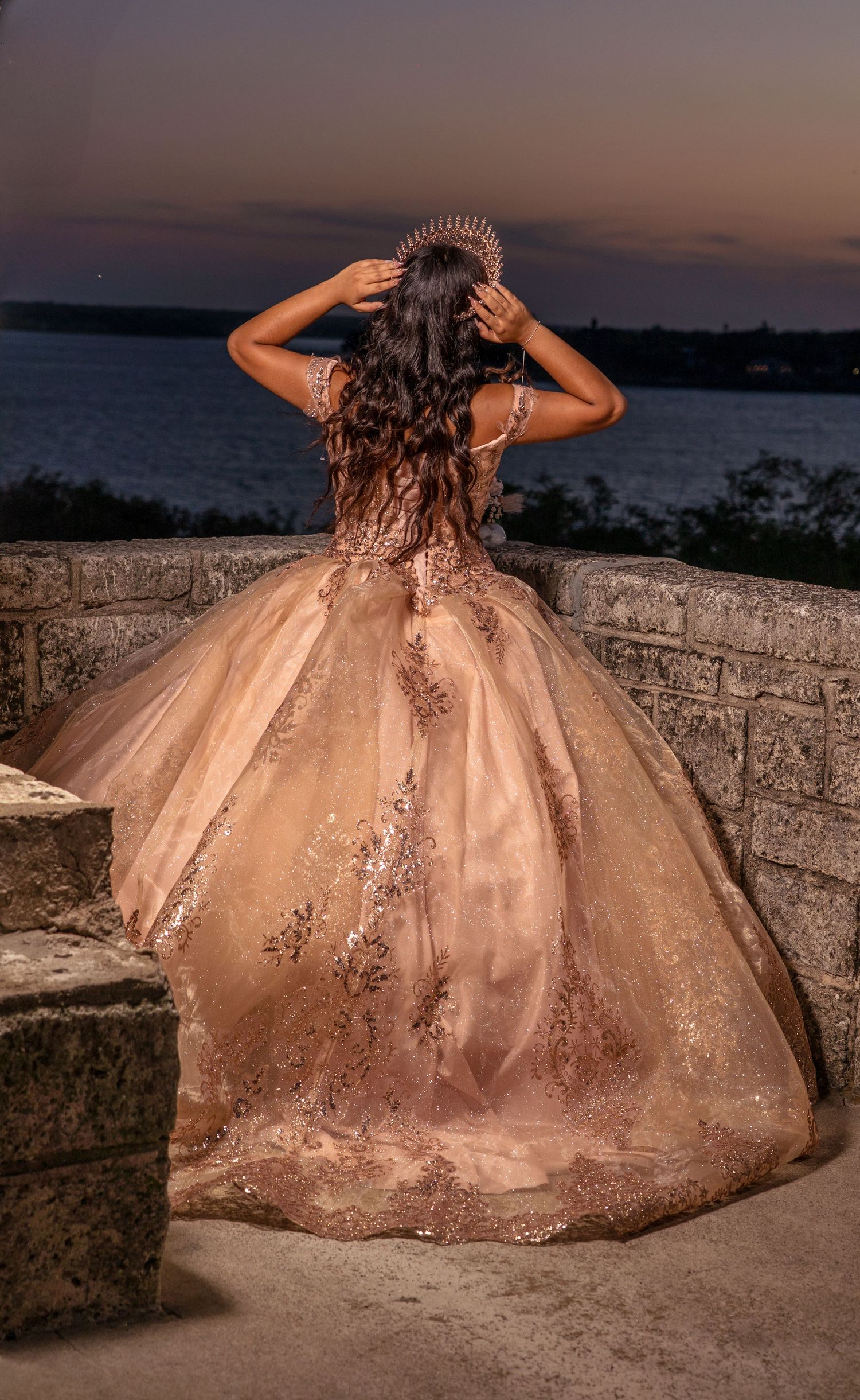 Sunset silhouette of sparkly ball gown overlooking waterfront from stone balcony.