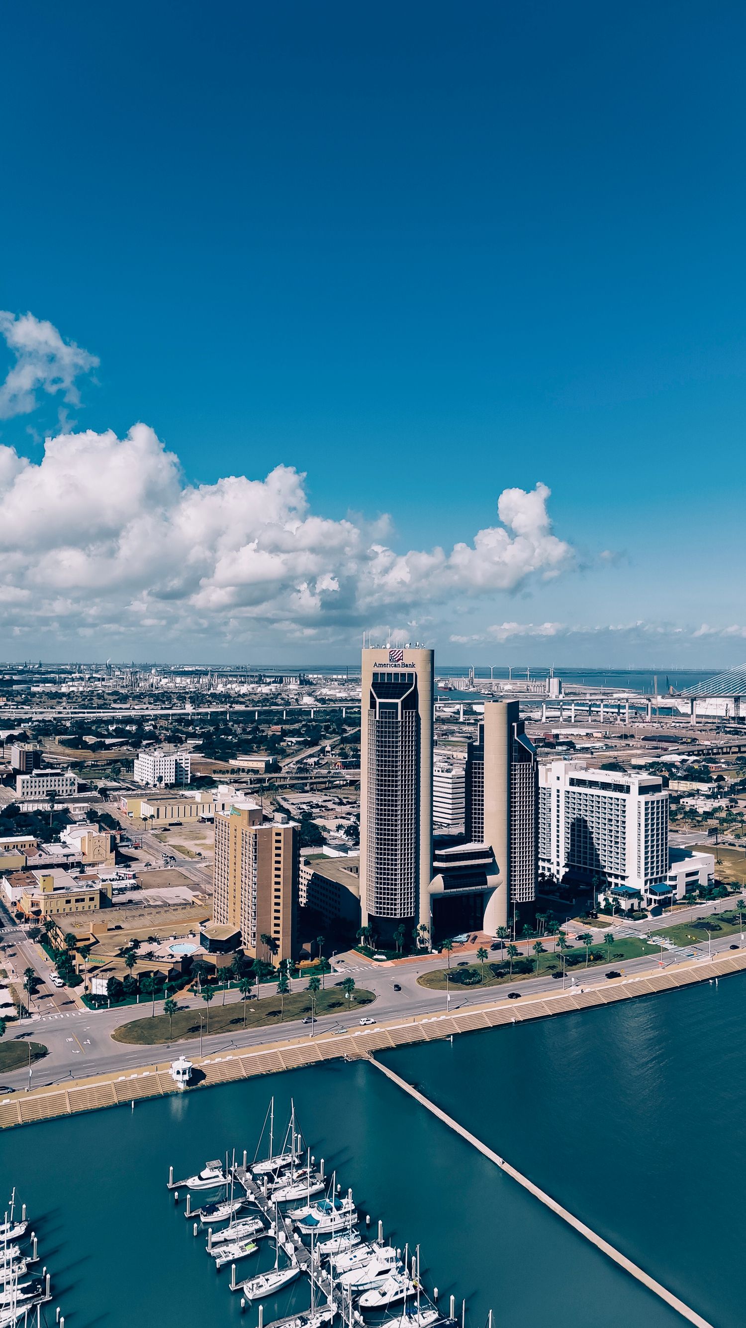 Aerial view of Corpus Christi skyline and marina with luxury boats under bright blue sky with white fluffy clouds.