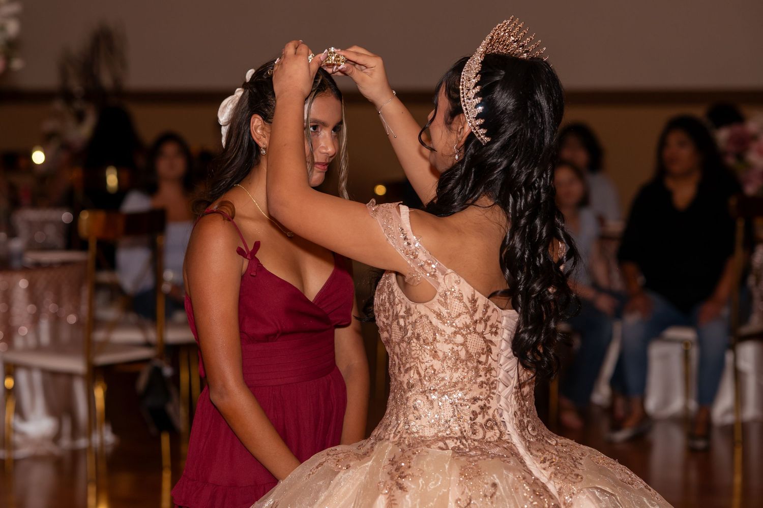 A quinceañera girl in a rose gold gown has her tiara placed by a friend in a burgundy dress at a celebration.