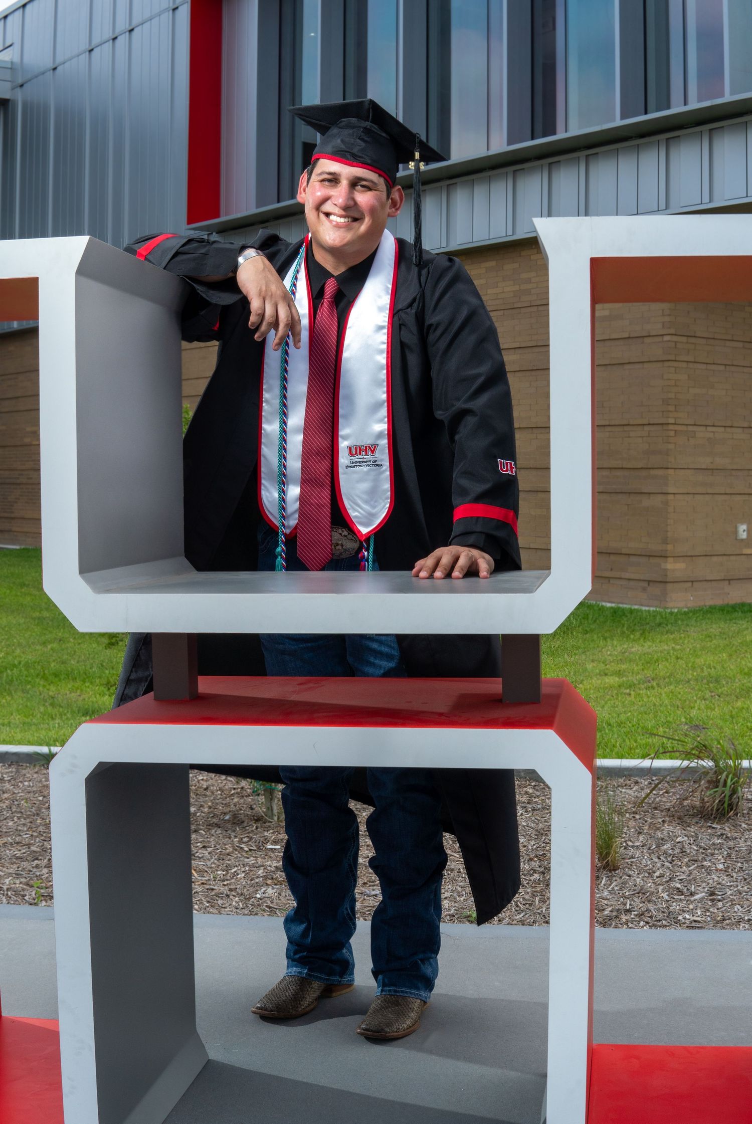 Graduate in academic regalia poses inside modern architectural display on campus grounds.