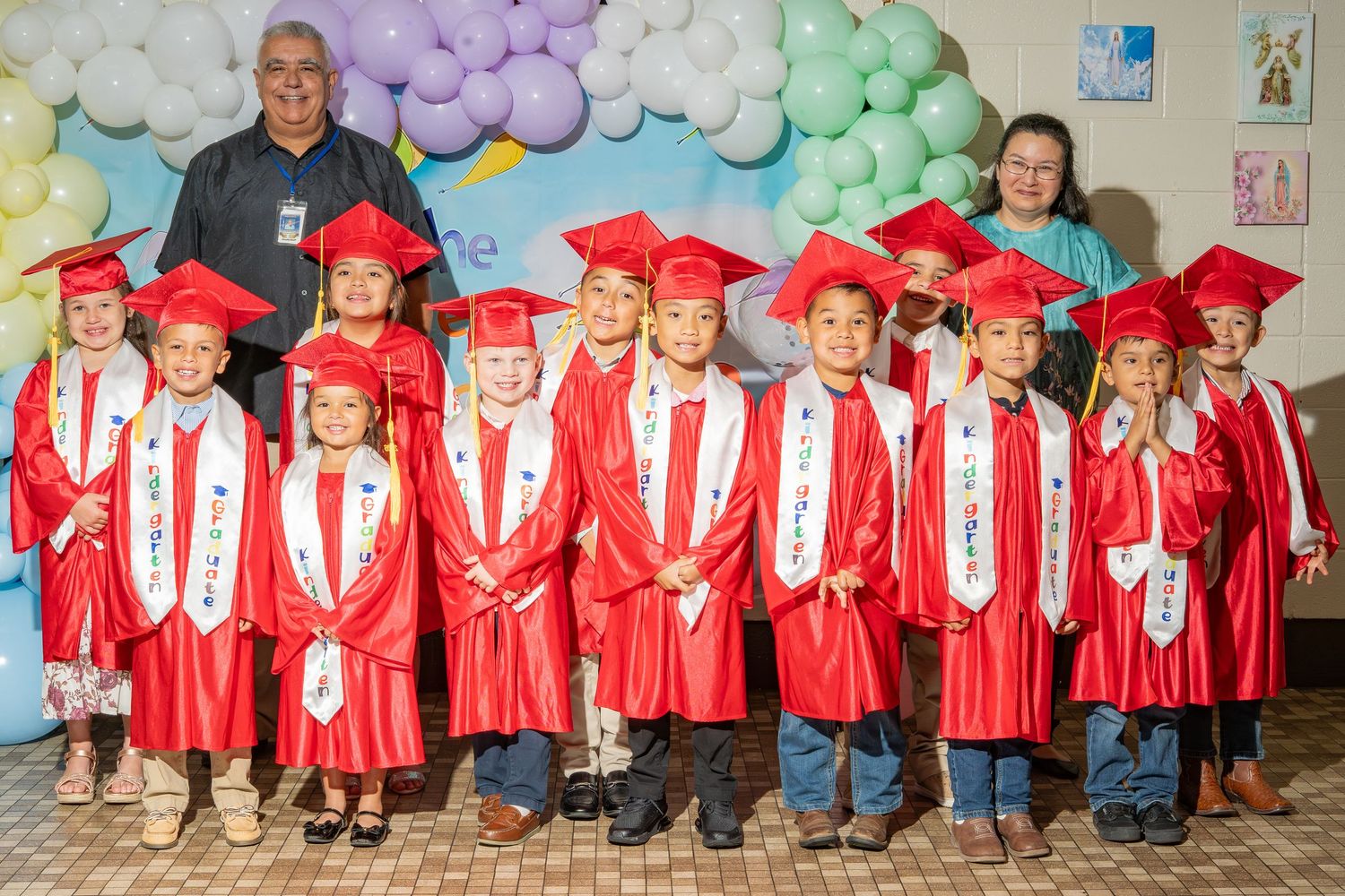 Young children in red graduation gowns and caps pose with two adults at a colorful preschool graduation ceremony.