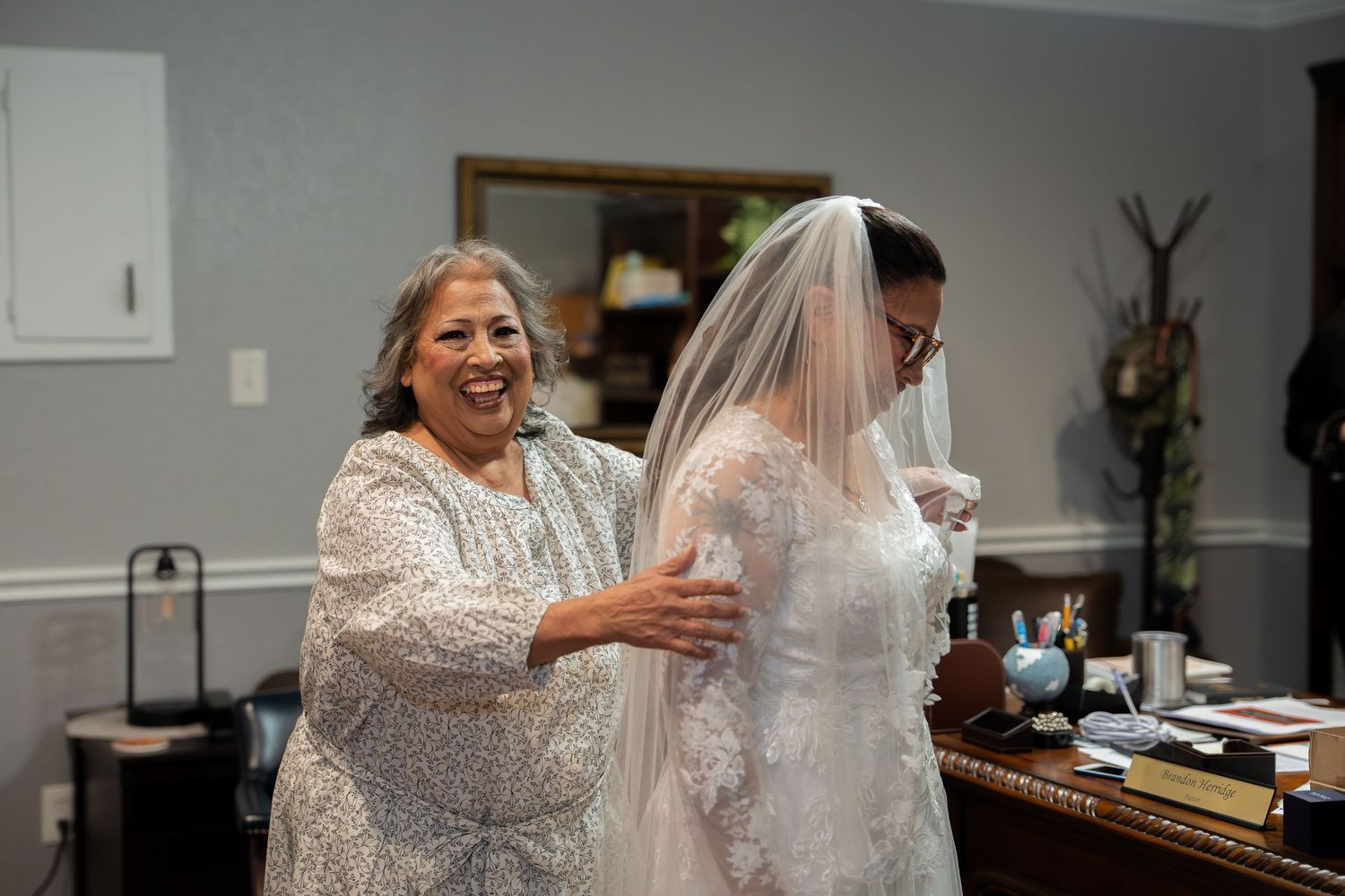 Smiling mother helps bride in lace wedding gown and veil get ready before the ceremony.