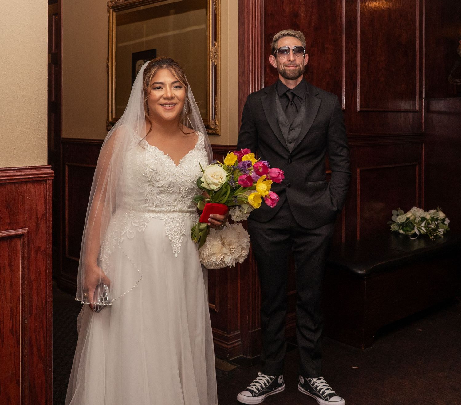 Bride in white lace gown with veil and groom in black suit holding colorful bouquet pose together at wedding venue.