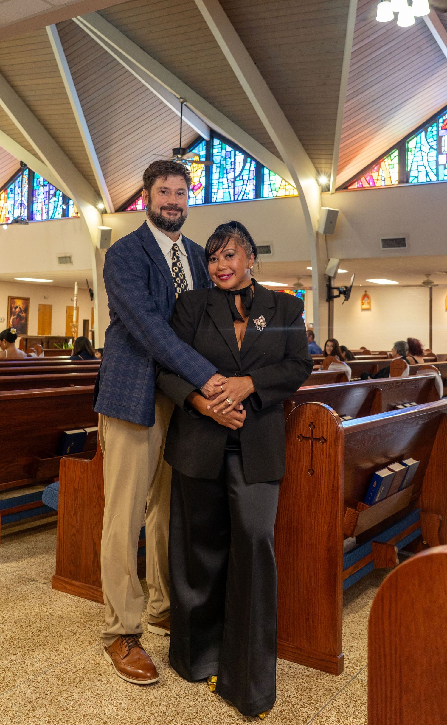 A couple stands together smiling in a church sanctuary with stained glass windows and wooden pews.