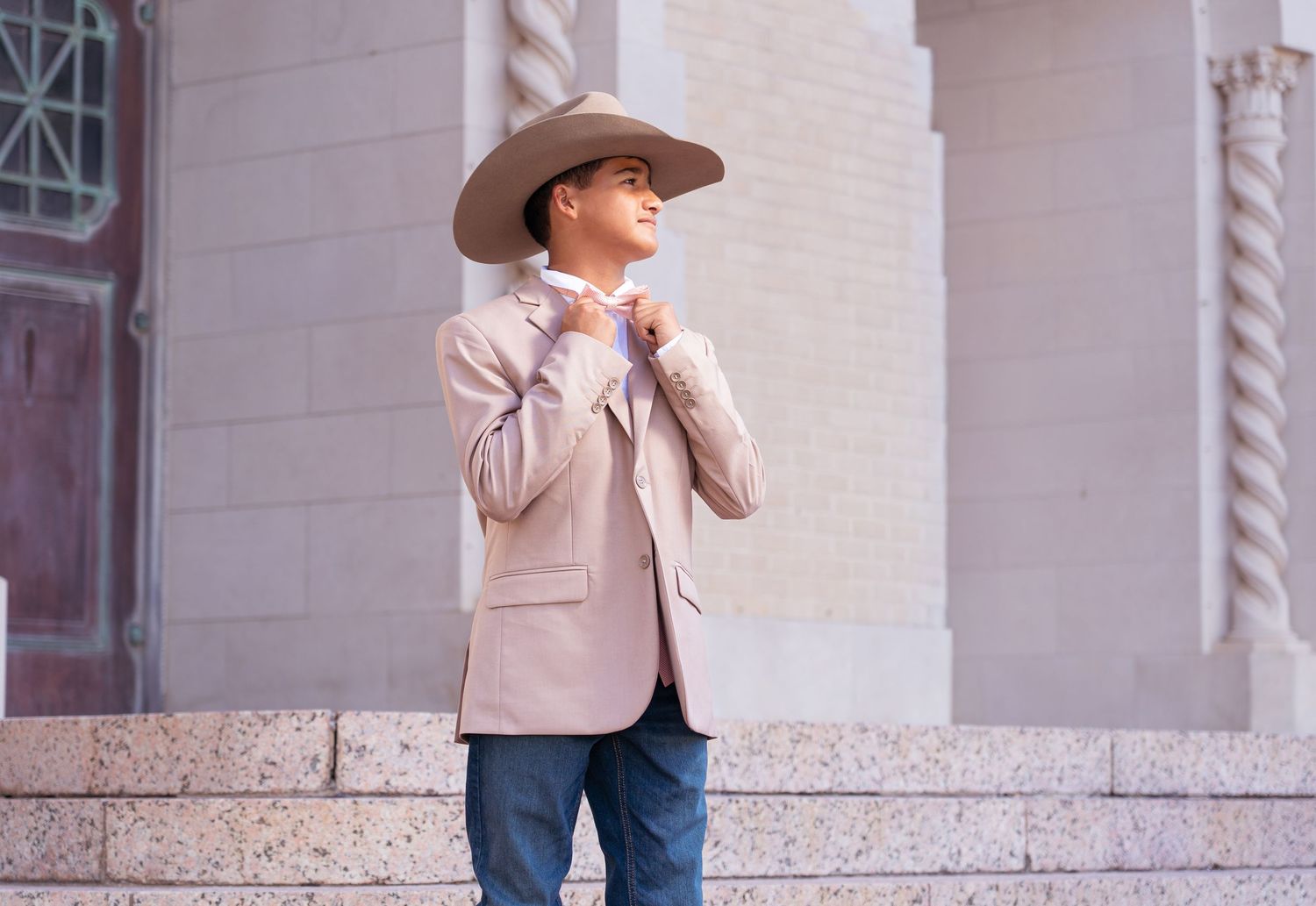 Person in beige blazer and cowboy hat stands against classical building facade with arched windows.