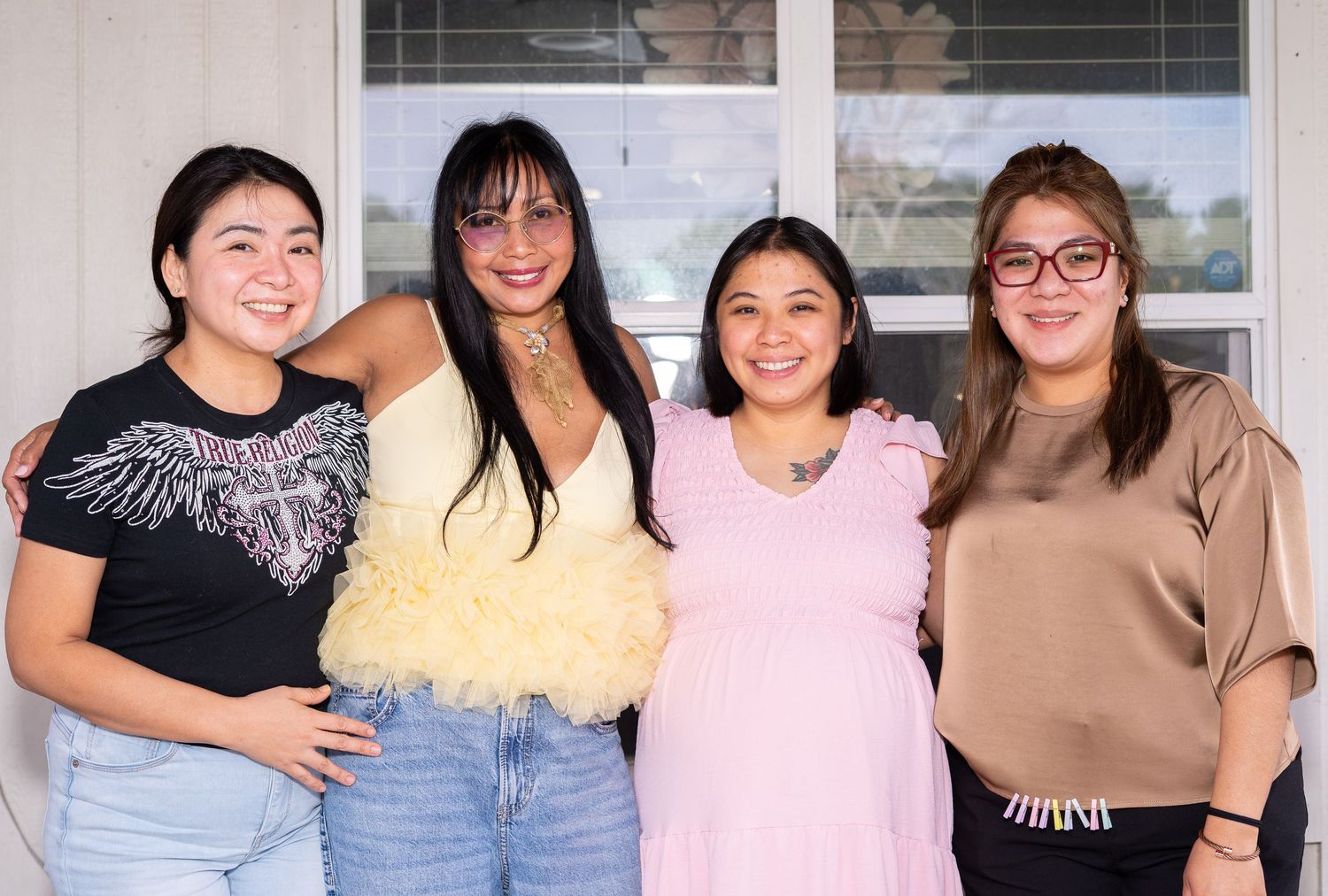 Four women smiling and posing together indoors, dressed in casual outfits ranging from black to pink and tan tops.