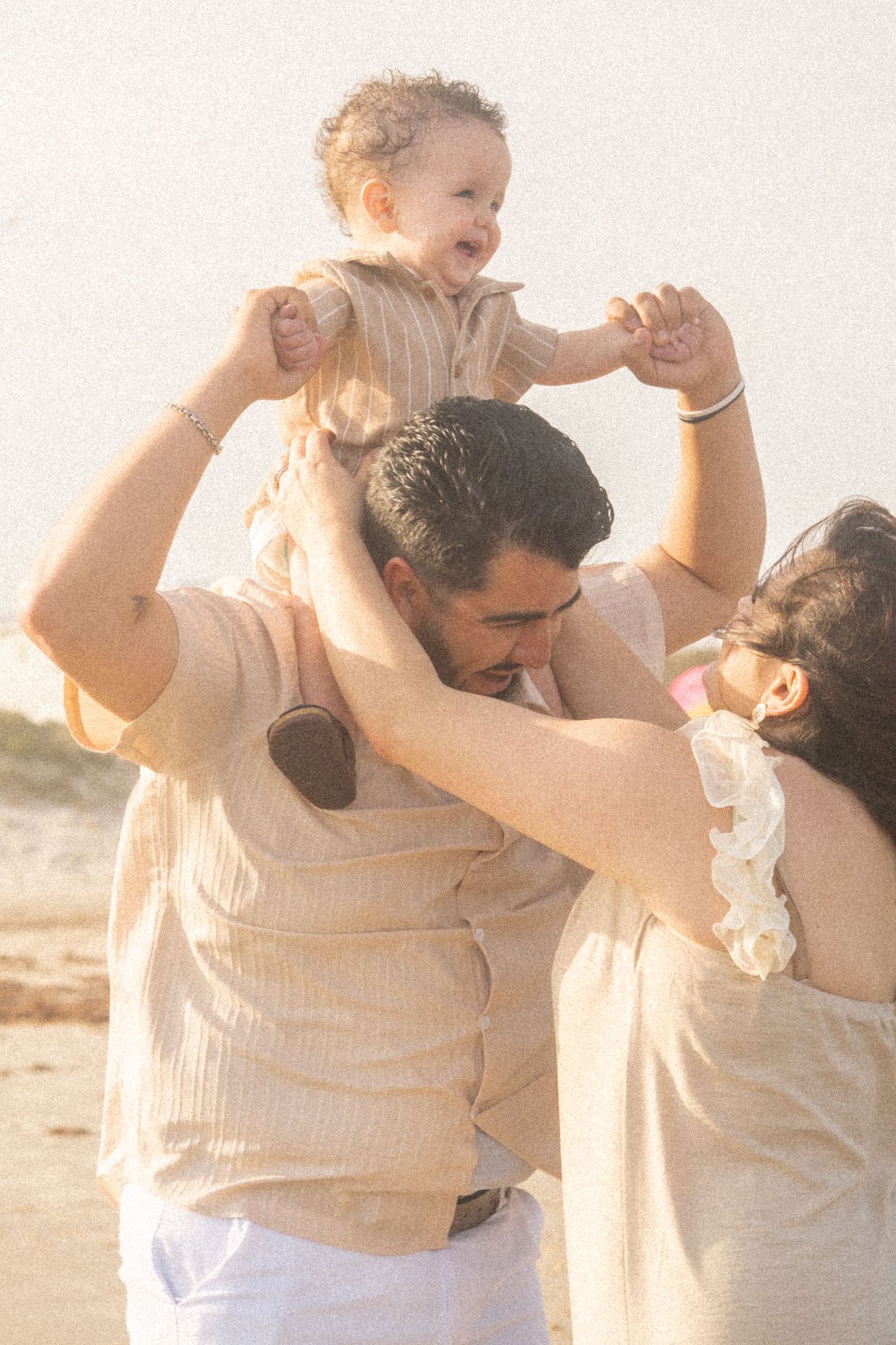 Happy baby sitting on dad's shoulders while mom helps hold hands during a sunny beach family photo.