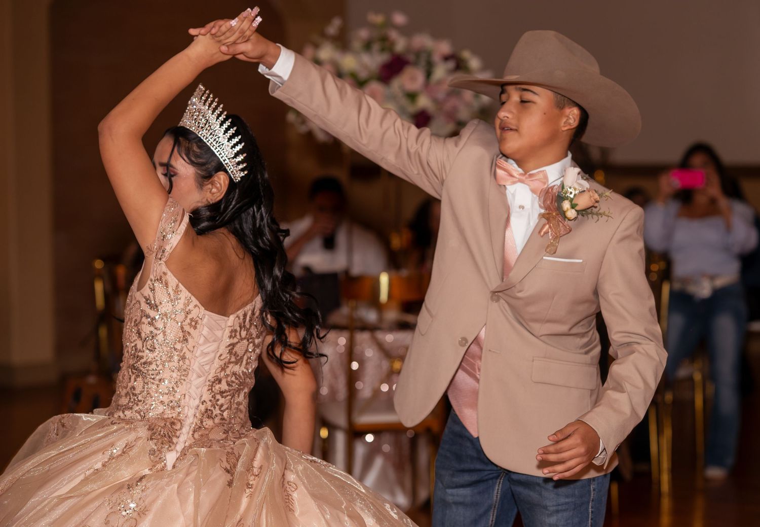 A young woman in a gold quinceañera gown dances with a partner in a tan cowboy hat.