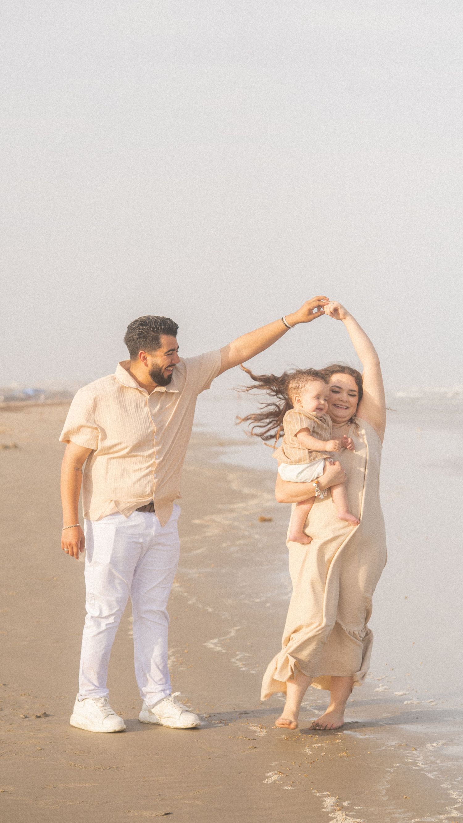 A joyful family with a baby dances together on a sandy beach, with the man twirling the woman by the hand.
