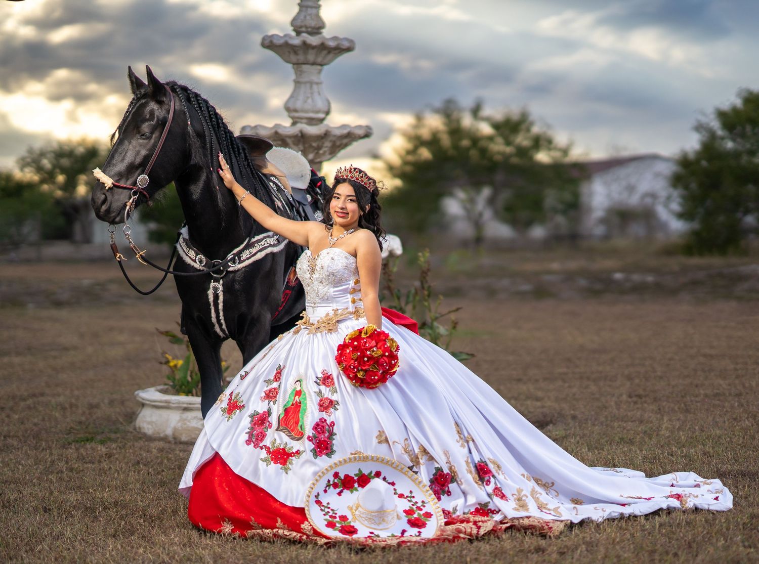 A black horse and someone in a white and red dress pose dramatically at sunset near a stone fountain.