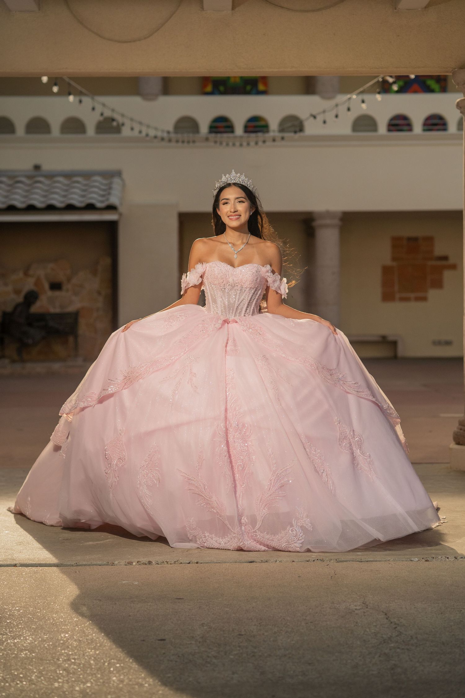 Young woman in a pink ball gown and tiara poses gracefully at her quinceañera celebration outdoors.