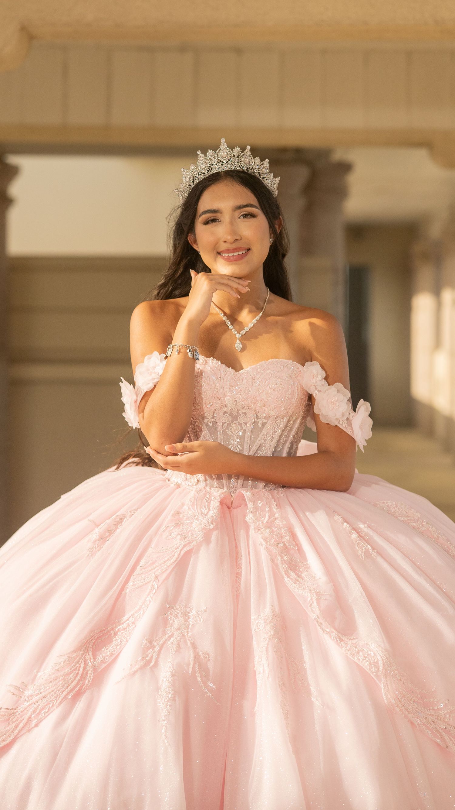Young woman in pink ball gown and silver tiara poses gracefully for quinceañera portrait outdoors.