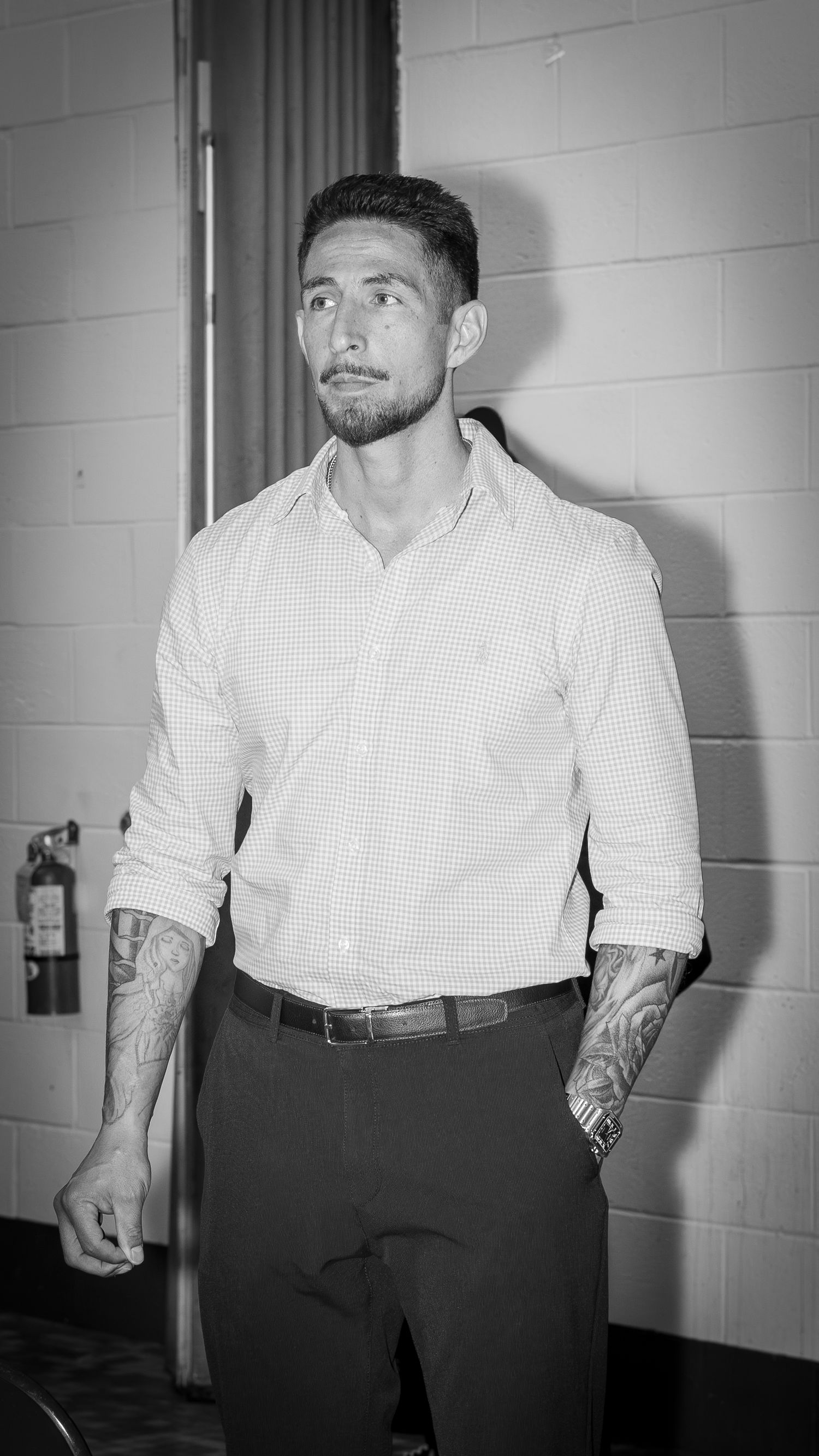 Black and white photo of a tattooed man in a button-up shirt and dress pants leaning against a brick wall.
